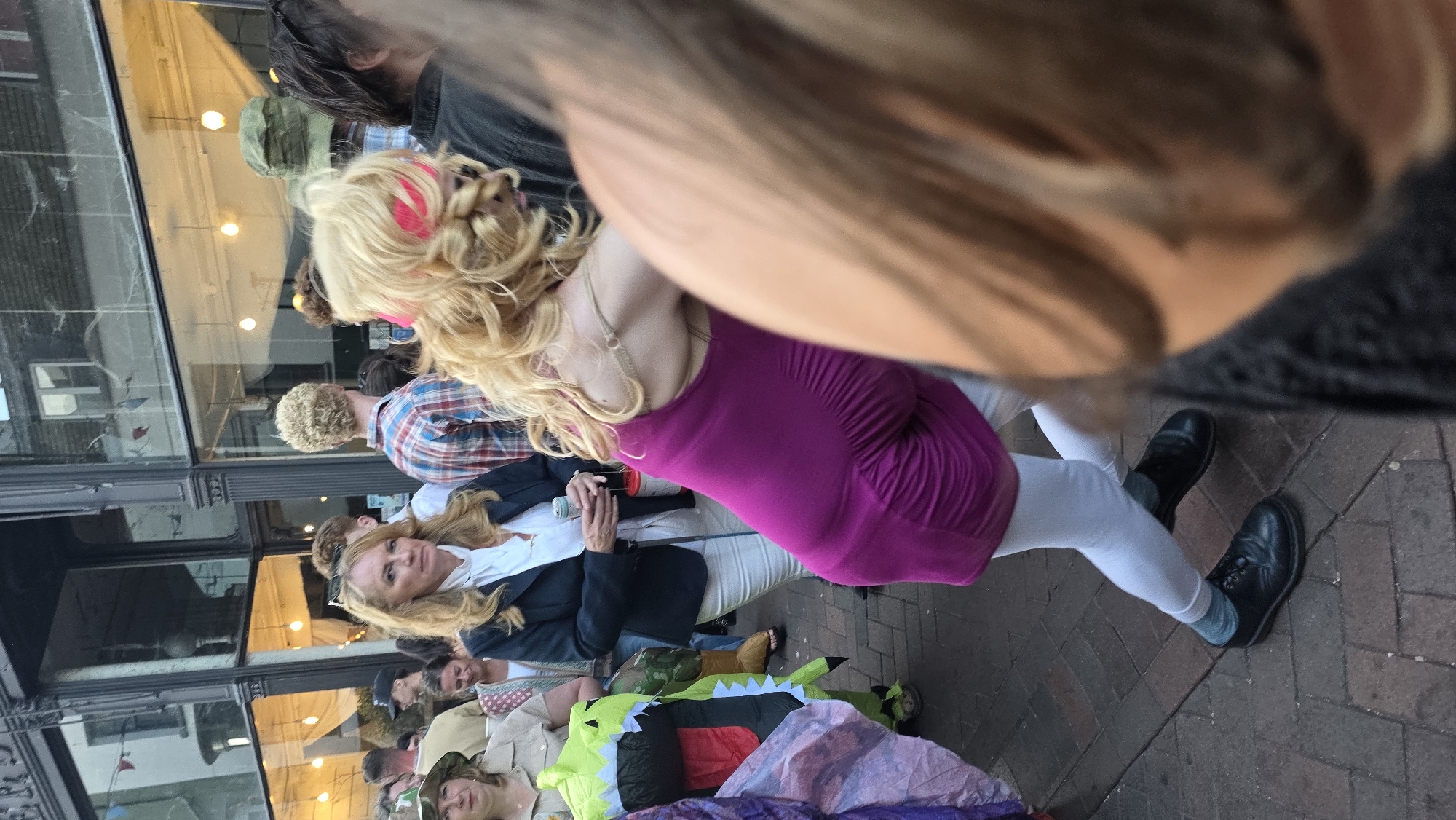 The image depicts a street scene in St Leonards, United Kingdom, likely during an event. In the foreground, a woman with blonde, curly hair and a pink headband is captured from the back, wearing a bright magenta, ruched dress and white leggings. She is wearing black lace-up shoes and grey socks. Her pose suggests she is either dancing or in motion. To her left, a woman with blonde hair and a white blouse under a dark blazer holds a can and a cup. She is looking towards the camera. Behind her, a man in a red and blue plaid shirt is visible. Further left, a person in a green dinosaur costume stands with their hands inside the costume. The background shows a building with large windows, lit from within, and a crowd of people on a brick-paved street. The overall atmosphere appears lively and informal.