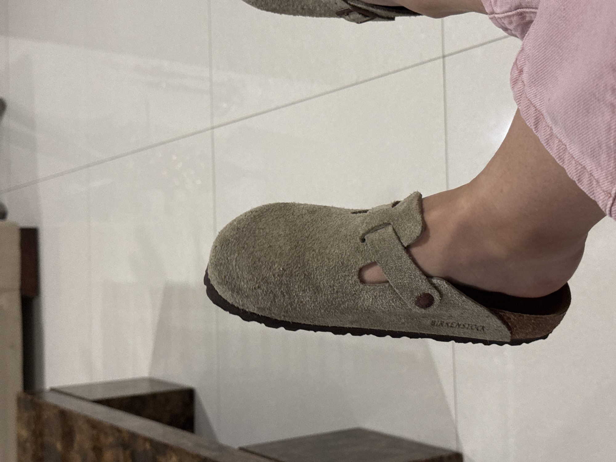 A person's foot is seen wearing a light brown suede Birkenstock Boston clog. The shoe has a brown cork sole and a black rubber outsole. The person is wearing pink denim pants. The background is a wall of white tiles. A dark wooden chair is partially visible in the bottom left corner of the image. The setting appears to be indoors. The city of Santa Clara Ocoyucan, Mexico is not visible in the image.