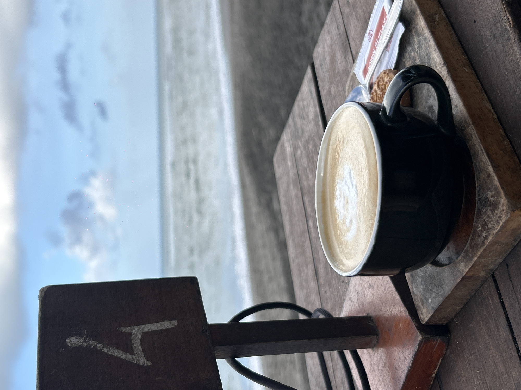 The image shows a latte in a black mug, resting on a square wooden coaster atop a weathered wooden table. Packets of brown sugar and a small cookie are also on the table. The background features a blurred view of a beach with gray sand, the ocean, and a cloudy sky. A dark wooden sign with a white symbol, possibly a logo, is partially visible in the lower left corner. The location is likely near a beach in Mengwi, Indonesia.