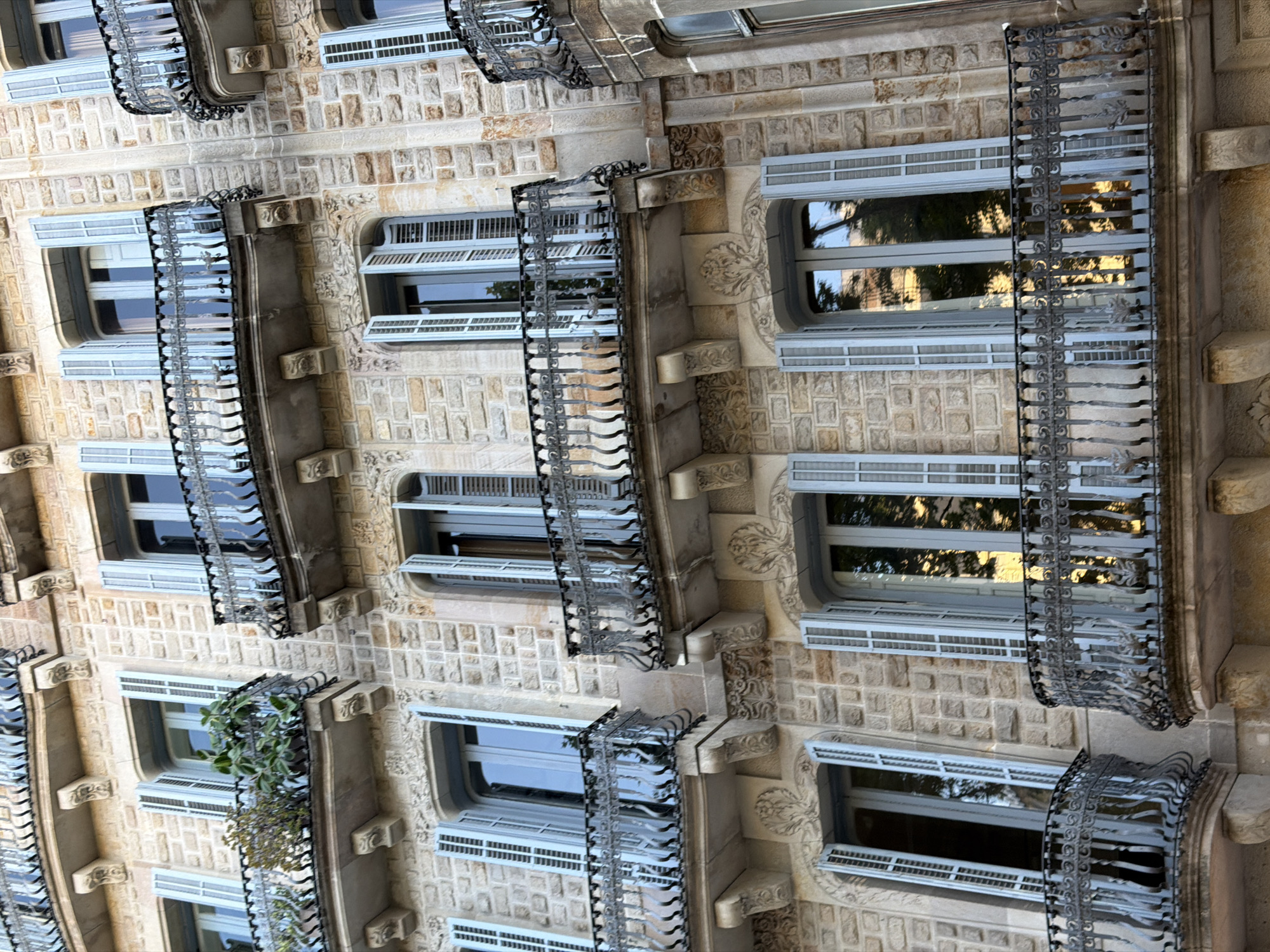 This media file features a close-up, low-angle view of the exterior of a building in Barcelona, Spain. The building's facade is constructed from light brown stone blocks, and it is adorned with ornate decorative elements, including carved stone reliefs and intricate wrought-iron balconies. Multiple windows are visible, many with light blue shutters that are partially open, revealing the dark interiors. The windows themselves appear to be modern, with multiple panes. Reflections of trees and sky are visible in the glass of some windows, suggesting it is daytime and possibly sunny. Some of the balconies have plants growing on them, adding a touch of greenery. The architectural style suggests an older, possibly historic building with some modern renovations. There is no visible text in the image. The scene is static, focusing solely on the architectural details of the building.