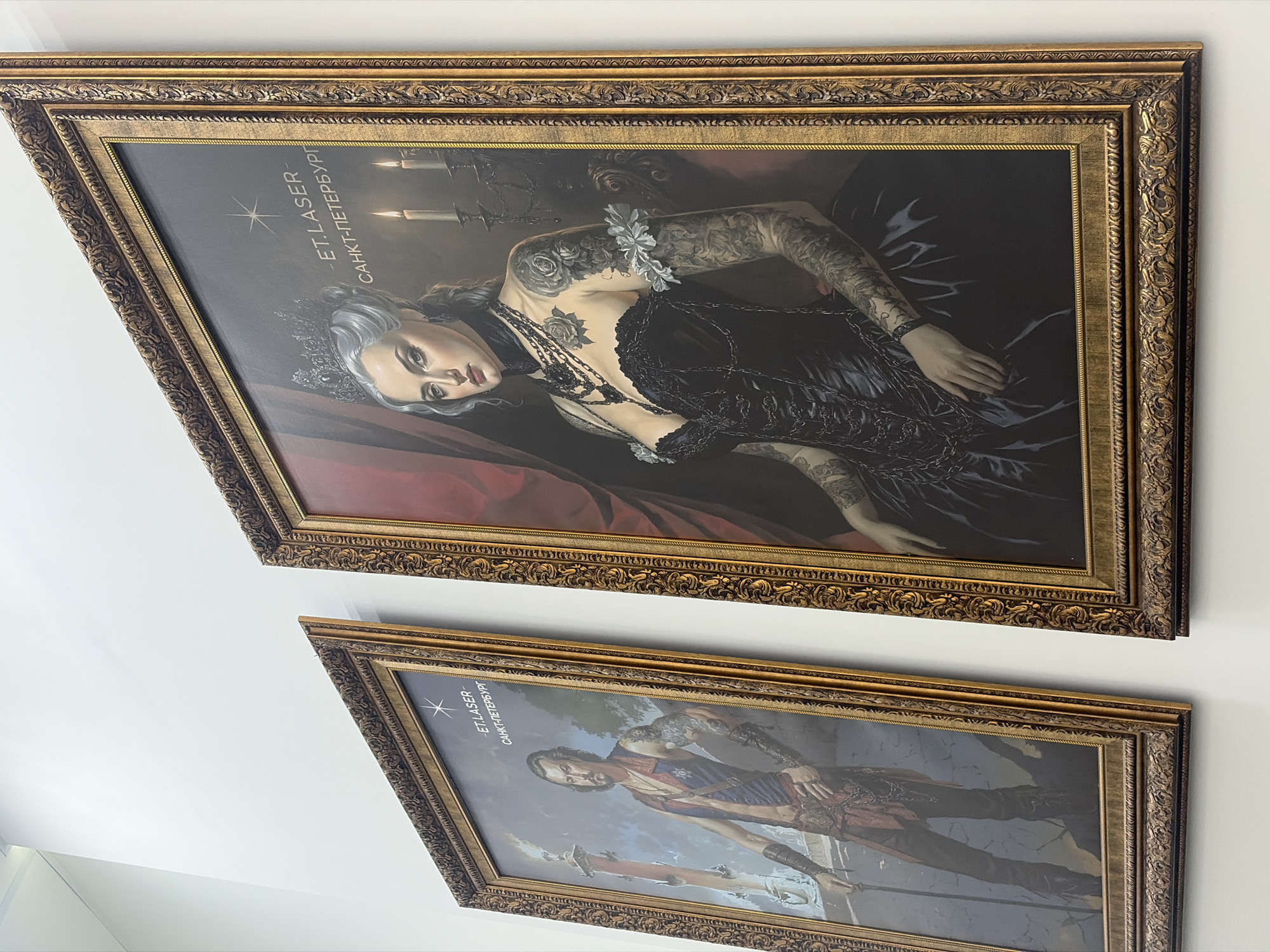 The media file showcases two large, ornately framed pictures displayed on a light-colored wall, likely indoors. The images are captured from an angled perspective, showing them slightly rotated.
The top picture is a portrait of a woman with light-colored (possibly white or light grey) hair, adorned with a crown and a dark, elaborate gown featuring chains. She has extensive black tattoos on her arms and chest. She is depicted in a reclining position, gazing directly at the viewer. Lit candles are visible in the dark background, contributing to a dramatic atmosphere. In the upper left corner, vertically oriented text reads: a star icon, "ET.LASER -", and "САНКТ-ПЕТЕРБУРГ" (Saint Petersburg).
The bottom picture, also set in an elaborate golden frame, presents a full-body portrait of a man. He has dark hair, a beard, and numerous tattoos covering his arms and chest. He is dressed in a historical or fantasy-inspired costume, including a blue and red sleeveless top, dark trousers, and arm guards, holding two swords. He stands facing the viewer. This artwork also features the same vertically oriented text in the upper left: a star icon, "ET.LASER -", and "САНКТ-ПЕТЕРБУРГ".
Both artworks feature heavily tattooed individuals, blending classical portrait aesthetics with modern body art, hinting at a gothic or fantasy theme. The opulent golden frames are intricately designed, enhancing the traditional art style. The visible text "ET.LASER -" and "САНКТ-ПЕТЕРБУРГ" suggests these pieces are displayed in a commercial establishment, likely a laser treatment clinic specializing in tattoos, located in Saint Petersburg, Russia.