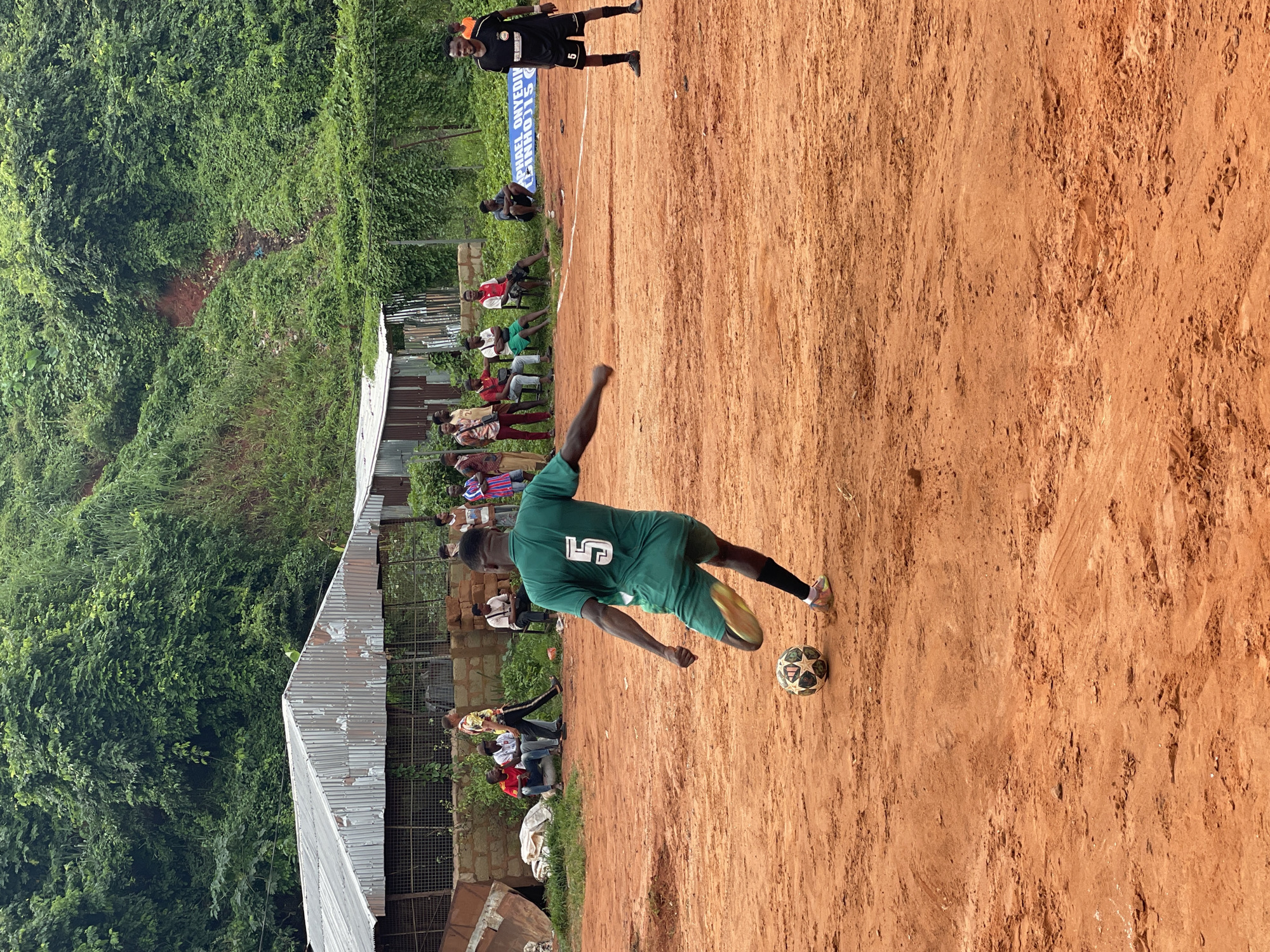 In an outdoor, dirt field with a lush green hill in the background, a group of young men are playing soccer. A player in a green jersey with the number 5 on the back is mid-kick, about to strike a soccer ball. Several other young men are scattered around the field, either watching the game or waiting to play. In the background, a makeshift shelter with a corrugated metal roof stands. The overall scene depicts a casual soccer game taking place in a community setting.