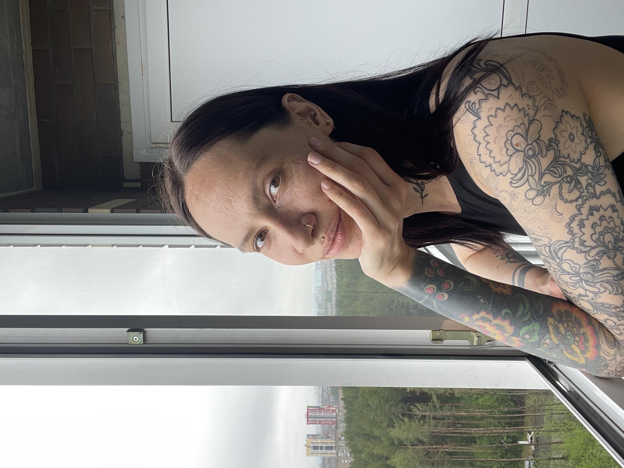 A young woman with tattoos is leaning on a window frame, gazing at the camera. Her dark hair cascades over her shoulder and arm, which is adorned with a vibrant floral tattoo. She has a nose piercing, and her face is framed by her hand. Outside the window is a cityscape with trees and a few high-rise buildings; the city appears to be Izhevsk, Russia. The weather outside appears overcast.