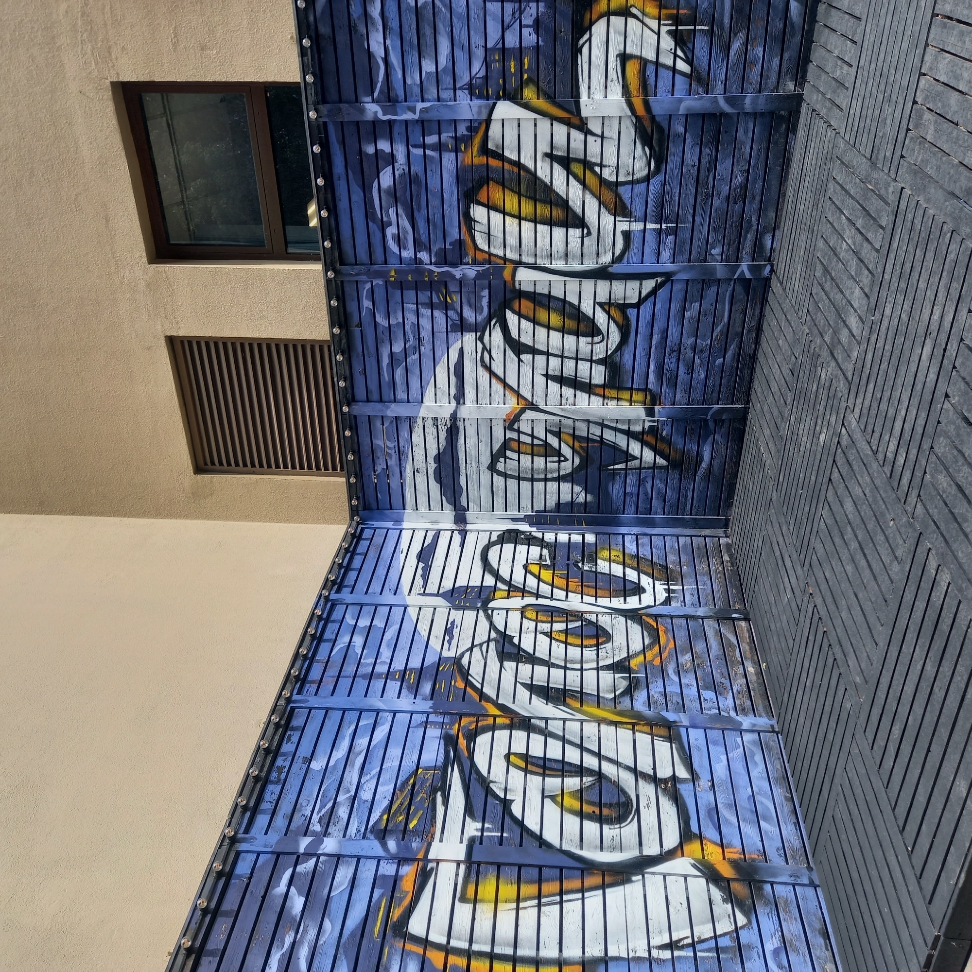 This image captures an outdoor corner area in Zvenigorod, Russia, featuring graffiti on blue slatted panels, a dark slatted floor, and a building wall in the background.
The foreground is dominated by a dark grey, slatted floor composed of interlocking square tiles arranged in alternating perpendicular directions. The slats are narrow and uniformly spaced.
Behind the floor, two sections of a vibrant blue slatted fence or wall meet at a 90-degree angle, forming an interior corner. The blue slats exhibit a worn, weathered appearance with areas of lighter blue or faded paint. Along the entire top edge of this blue structure, a row of small, clear, spherical lights is uniformly spaced, mounted on a dark horizontal rail.
The blue slatted surface is adorned with large, stylized graffiti lettering. The letters are primarily white with an interior gradient of yellow and orange, outlined in black. The graffiti appears to be composed of two distinct word segments, separated by the corner of the structure. The left segment, on the left panel, features letters that visually resemble "GAROC" (G, A, R, O, C, or similarly stylized forms). The right segment, on the right panel, displays letters that visually resemble "GALZ" or "GAJZ" or a stylized Cyrillic "GAЖ". The graffiti extends across the horizontal slats, giving it a somewhat striped appearance due to the gaps. A subtle, lighter blue abstract design, possibly representing clouds or sky, is painted on the blue slats behind the main graffiti letters.
In the upper background, a flat, light beige or tan stucco-textured building wall rises above the blue structure. On this wall, to the left of the corner, there is a rectangular dark brown louvered vent. To the right of the corner and the vent, a rectangular window with a dark brown frame is visible. The window glass reflects its surroundings, appearing dark and showing faint outlines of what might be trees or buildings.
The scene is brightly illuminated by natural daylight, casting minimal shadows and indicating an open, outdoor setting. No people or overt actions are depicted.