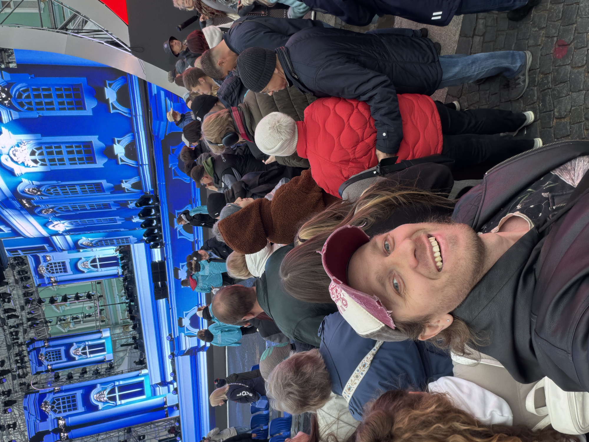 A man is taking a selfie in a crowd of people, with a large screen in the background showing a projected image of the Winter Palace in Saint Petersburg, Russia. The crowd consists of adults of varying ages and dress, some wearing winter hats and coats. The man is wearing a baseball cap and a black jacket. The screen shows a blue and white facade with arched windows. The location appears to be an outdoor event or gathering. The ground seems to be paved with cobblestone.