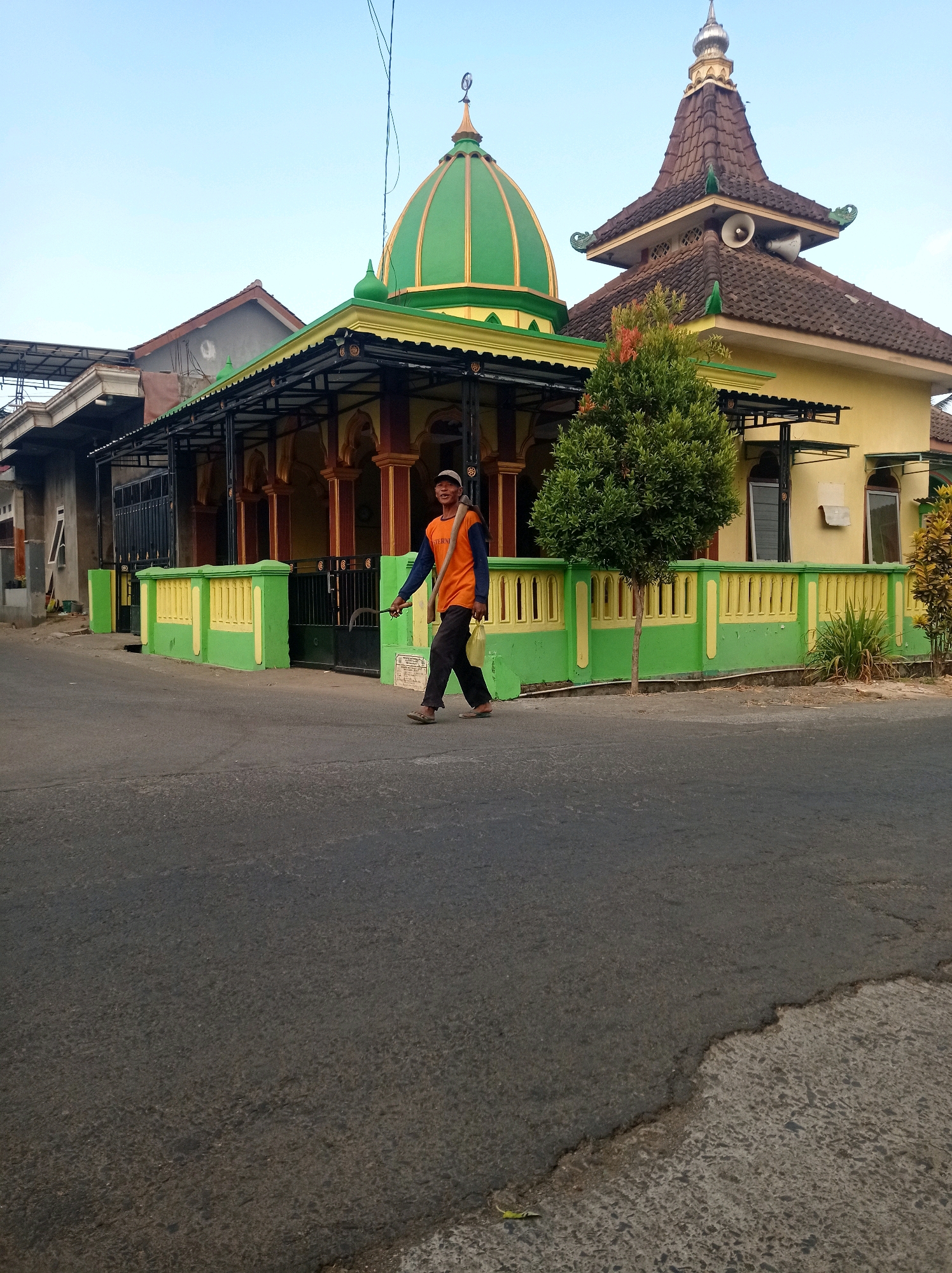 In the city of Purwosekar, Indonesia, a man walks across an asphalt road in front of a building with green and yellow accents. The building appears to be a mosque, evidenced by its green dome with gold trim and a smaller, darker tiled tower with a speaker on it. The mosque has a covered walkway in front, supported by red and gold columns, and is bordered by a low, green and yellow fence. The man is wearing an orange long-sleeved shirt, dark pants, and sandals, and carries a tool with a wooden handle over his right shoulder, as well as a yellow bag in his left hand. He is walking from left to right, facing slightly towards the viewer and smiling. The sky is clear and blue. The surrounding environment suggests a residential or community area, with glimpses of other buildings and vegetation.