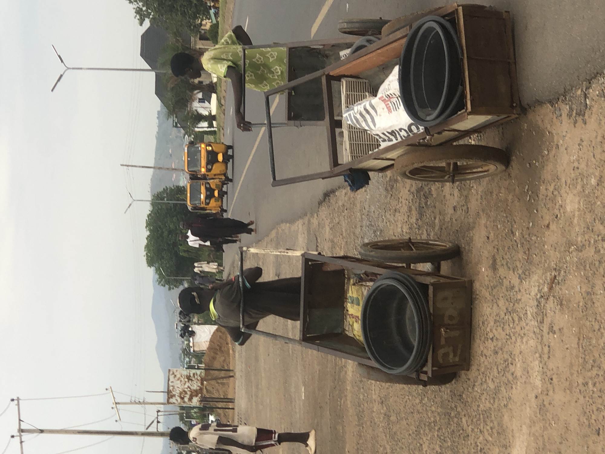 Two men are pulling hand-drawn carts on an asphalt road. Each cart carries large black circular items. Yellow three-wheeled vehicles are visible in the background. People are walking on the road. The location is in Abuja, Nigeria.