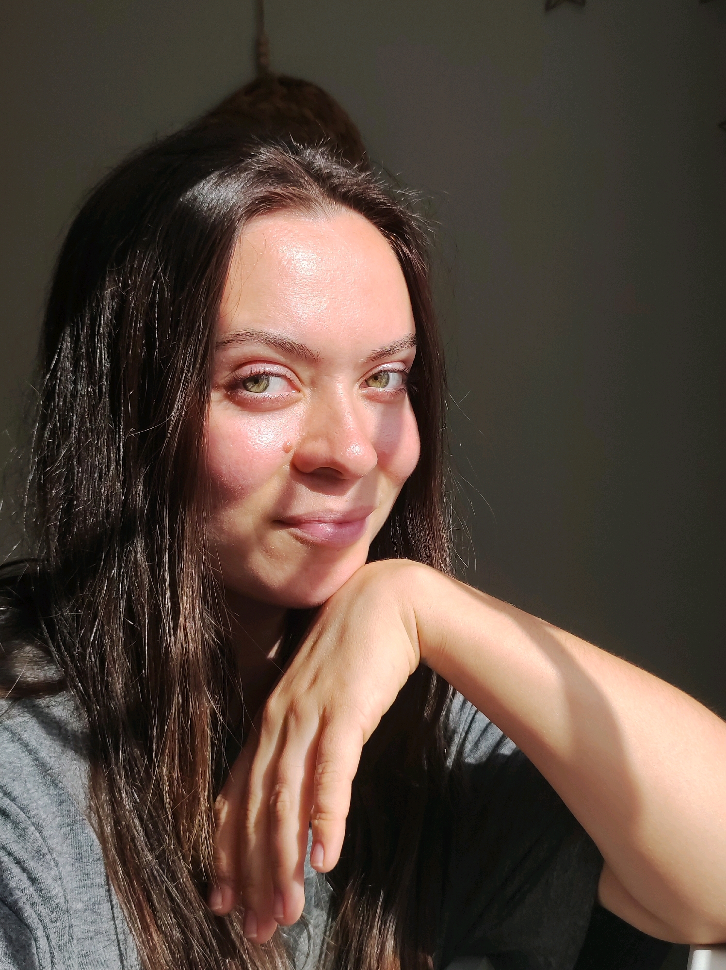 A close-up, selfie-style, head-and-shoulders portrait of a young woman with long, dark brown hair, possibly taken indoors with natural light. The woman is looking towards the camera with a slight smile, her right hand resting on her chin. Her left arm is visible in the foreground, with a stripe of sunlight across it. The background is dark and indistinct, with a hint of a wall and possibly some objects hanging from above. The lighting creates a dramatic effect, with strong highlights and shadows on the woman's face and arm. The woman is wearing a grey t-shirt. The image does not contain any information about the city of Chaikovsky, Russia.