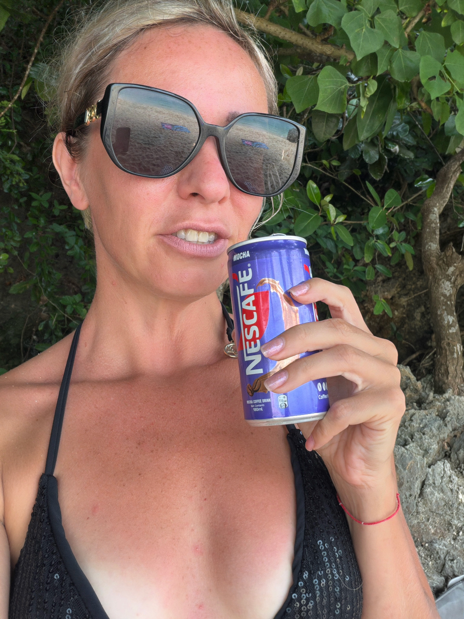 A woman with blonde hair pulled back and wearing large, dark sunglasses is holding a can of Nescafe Mocha coffee drink in her right hand. She is wearing a black sequined bikini top. The can is blue with red and white branding. Her left hand rests on her chest. The background shows green foliage and a tree trunk. The reflection in her sunglasses shows a sandy beach and some colorful items on the sand. Her skin appears tanned with some redness, suggesting sun exposure. There is a thin red string bracelet on her left wrist. The setting appears to be an outdoor, tropical environment, possibly near a beach. Based on the presence of tropical foliage and beach imagery in the sunglasses' reflection, this scene is consistent with a location like Malay, Philippines, known for its beaches.