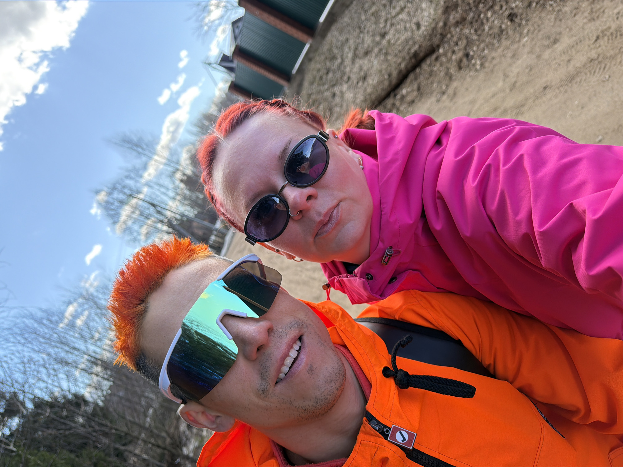 A selfie of two individuals. The person in the foreground, presumably taking the photo, has orange hair, a short cut, wearing sunglasses with white frames and mirrored lenses. This person is also wearing an orange jacket. Behind and slightly above this person is another individual. This second person has red hair styled in braids, sunglasses with black frames, and is wearing a pink jacket. The background shows what appears to be a path, possibly dirt or gravel, a fence line, some trees and a blue sky with white clouds. Based on information provided from the user, this image was taken in the city of Chaikovsky, Russia.