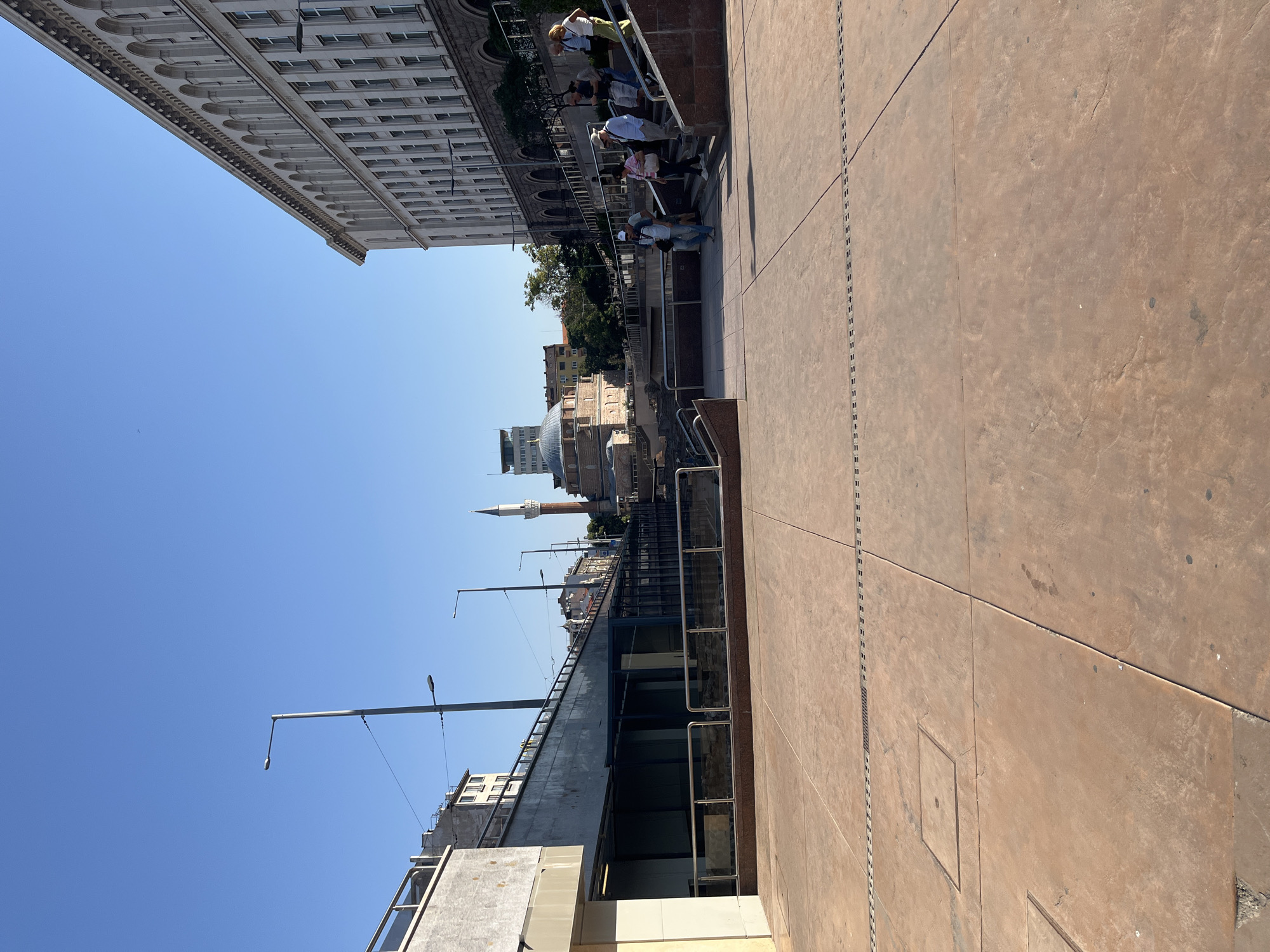 The image depicts an outdoor urban scene in Sofia, Bulgaria, likely near the Banya Bashi Mosque and the Serdica ancient complex. In the foreground, a large, reddish-brown tiled plaza with metal grates and access covers stretches across the bottom of the frame.
To the left, a modern structure with a gray concrete facade and glass barriers is visible, suggesting an entrance or exhibition space. Further back, a raised walkway or bridge with metal railings is present.
In the middle ground, a historic building with a dome and a tall, slender minaret is prominently featured. This is likely the Banya Bashi Mosque. Surrounding this building are remnants of ancient ruins, partially enclosed by glass and metal barriers. People are seen walking on stairs leading down towards these ruins.
To the right, a large, light-colored building with a long facade featuring numerous arched windows and decorative cornices dominates the background. This building appears to be an institutional or governmental structure. A group of individuals, dressed in casual attire, are walking up a set of wide, red-granite stairs leading to this building. They are interacting casually, with some looking around.
The sky is clear and blue, indicating a sunny day. Streetlights are visible throughout the scene, mounted on tall poles. The overall impression is a blend of historical and modern architecture within a public urban space.