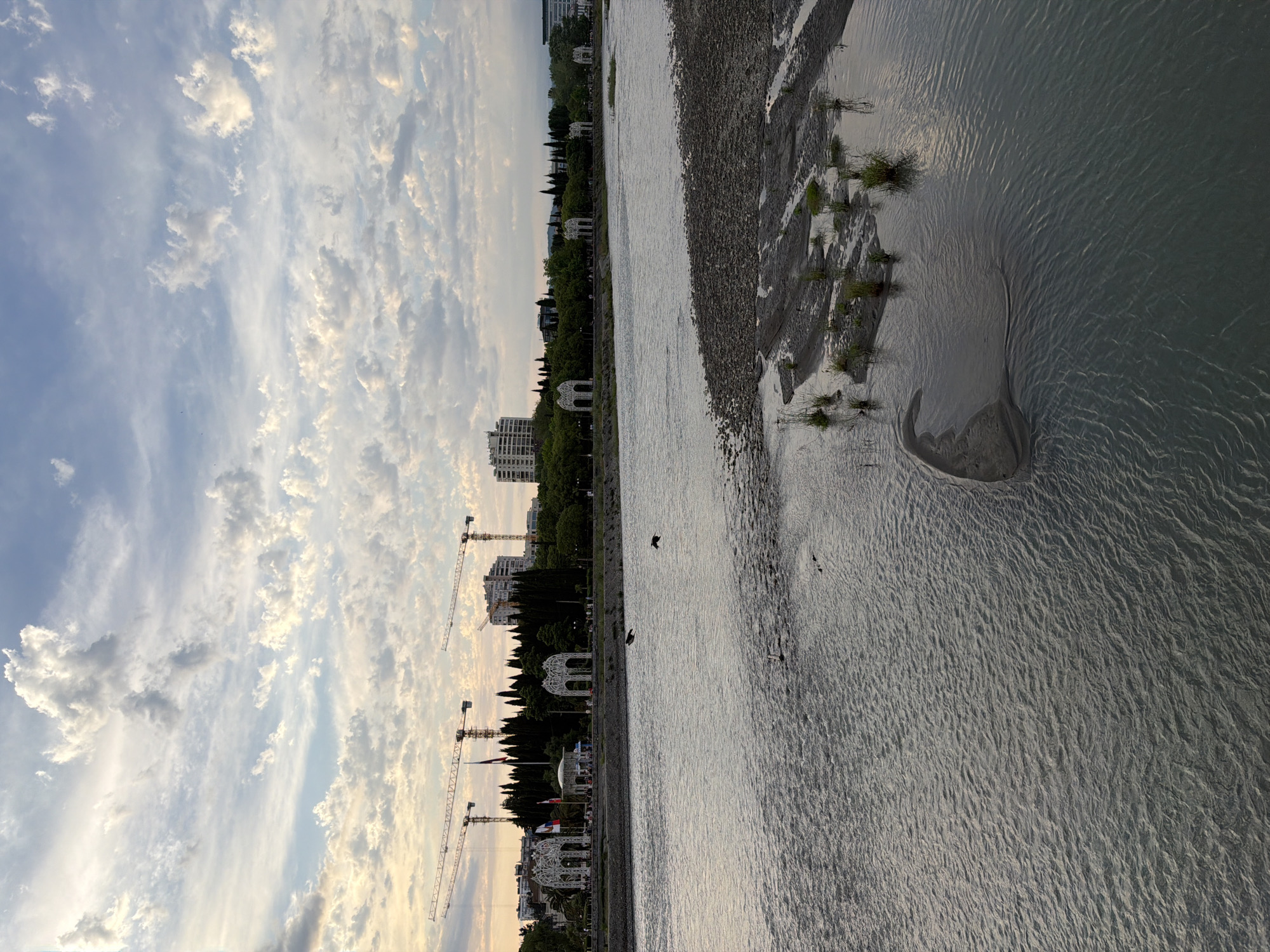 The image depicts a scenic view of a riverbank in Sochi, Russia, captured during the late afternoon or early evening, as indicated by the soft, warm light and the presence of long shadows. The sky is adorned with a dramatic display of scattered clouds, ranging from wispy cirrus to more substantial cumulus formations, catching the sunlight and creating a sense of depth and movement.
The foreground is dominated by the tranquil waters of the river, reflecting the sky and exhibiting subtle ripples and patterns on its surface. A sandy or pebbly embankment lines the river, with patches of hardy green grass emerging from the moist soil, suggesting a semi-arid or transitional environment.
Beyond the embankment, a lush green line of trees forms a natural border, interspersed with elegant white gazebos, hinting at a well-maintained public space or park. Further back, modern apartment buildings rise above the trees, their structures illuminated by the fading sunlight. The presence of several tall construction cranes along the coastline suggests ongoing development in the area. The scene is serene and peaceful, with no discernible human figures or specific activities, evoking a sense of calm and natural beauty. The time of day and the soft lighting contribute to a tranquil and picturesque atmosphere.