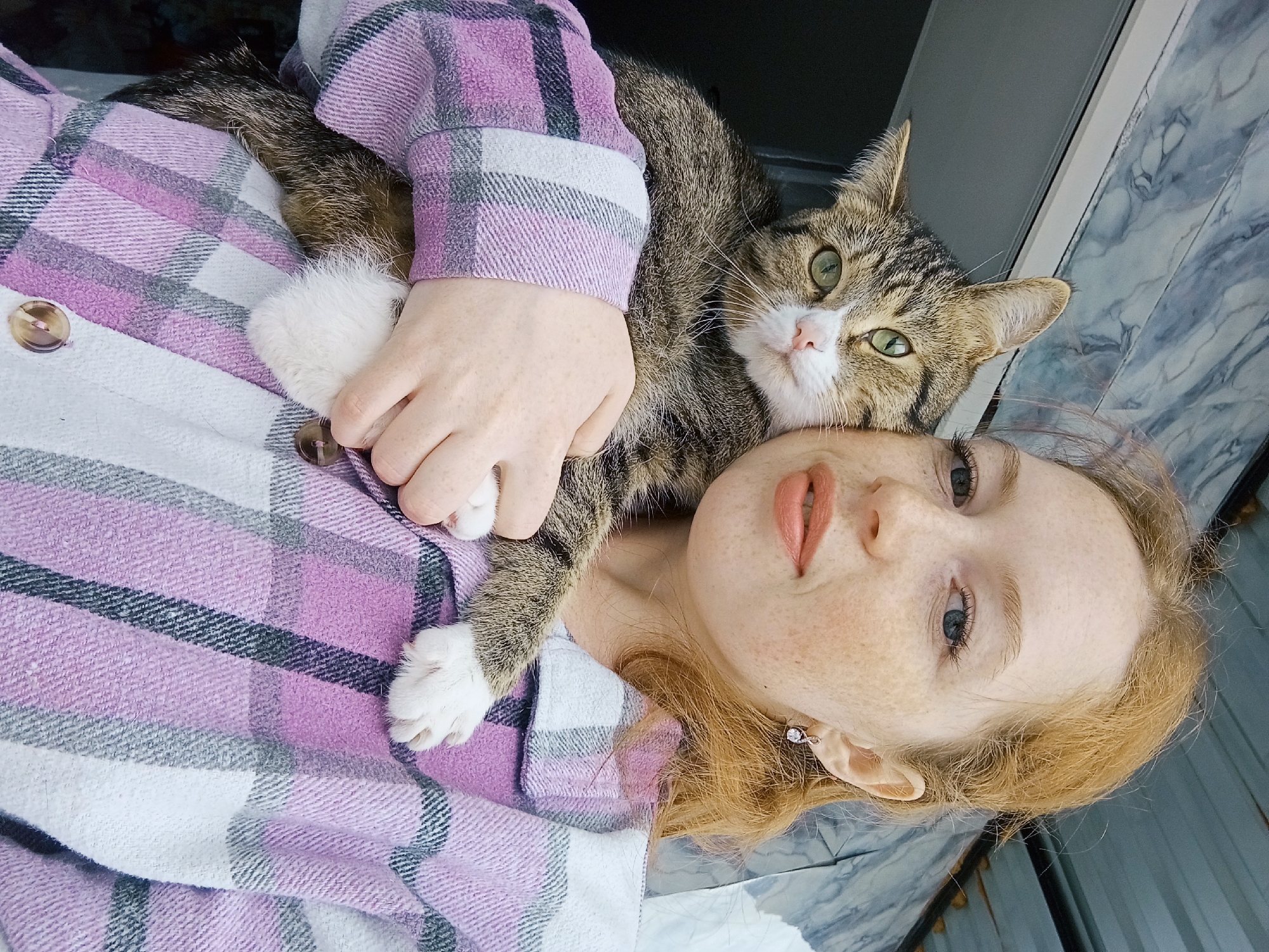 A fair-skinned woman with red hair and freckles lies on her back, cradling a brown tabby cat. The cat rests its head on her forehead. The woman is wearing a lavender and gray plaid shirt. The location is Yurgamysh, Russia. The background is a marble surface. The woman is looking at the camera. The cat has green eyes. One of the cat's white paws is resting on the woman's chest.