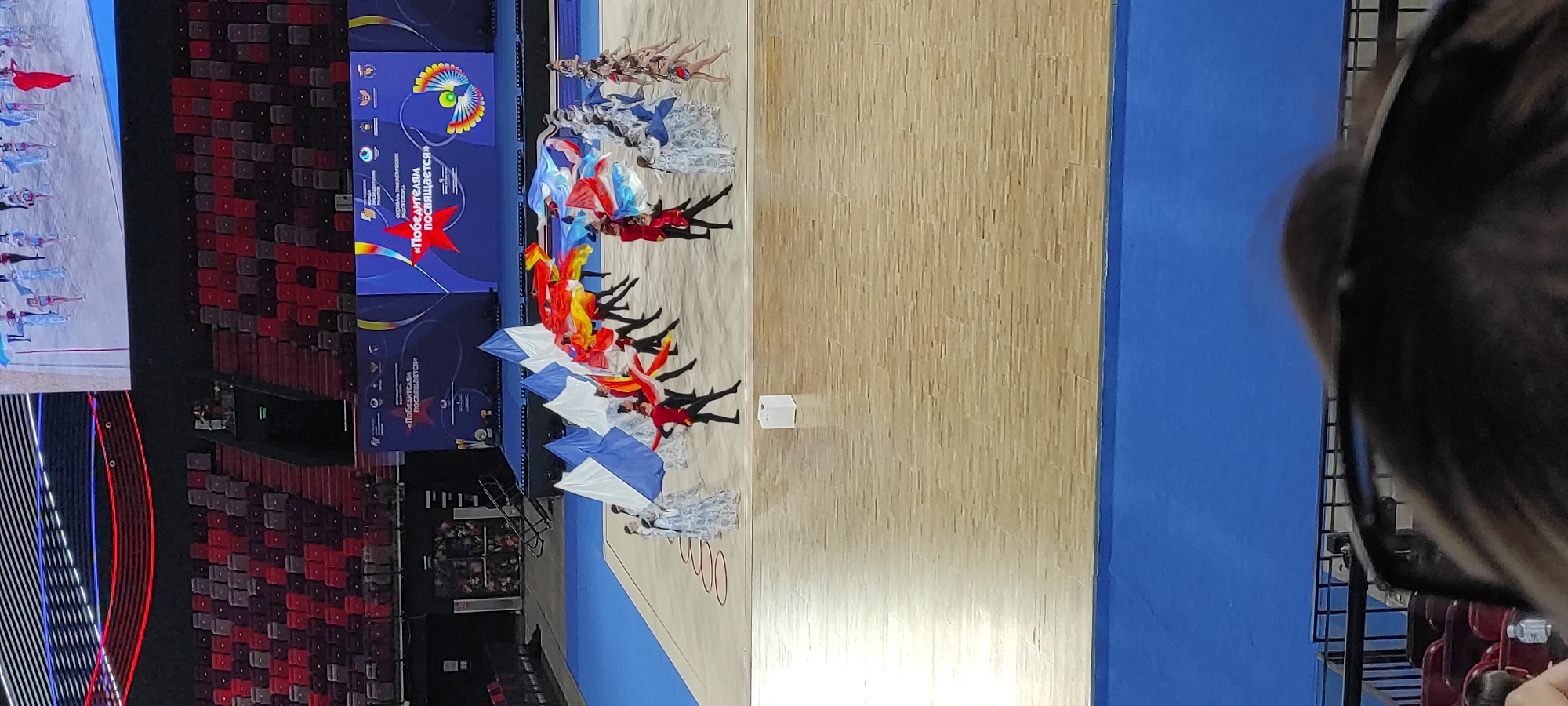 This image depicts a performance in a large indoor arena, likely a sports complex or concert hall. The floor is a polished wooden gymnasium floor, with a blue matting extending to the left. The primary focus is a group of performers on the floor, some holding large, flowing flags in red, white, and blue, suggesting a patriotic theme. These performers appear to be athletes or dancers, dressed in varying costumes, some in red and black, others in light blue and white. In the background, there are tiered seating areas filled with what appear to be spectators, though they are mostly out of focus. Above the seating, a large screen displays graphics, including text in Cyrillic that reads "ПОБЕДИТЕЛЯМ ПОСВЯЩАЕТСЯ" (Dedicated to the Winners) and a stylized red star. The overall atmosphere suggests a celebratory event, possibly an awards ceremony or an opening/closing ceremony for a sporting competition. Based on the context provided in other images in this set, this event is likely the Rhythmic Gymnastics World Championships, held in Moscow, Russia.