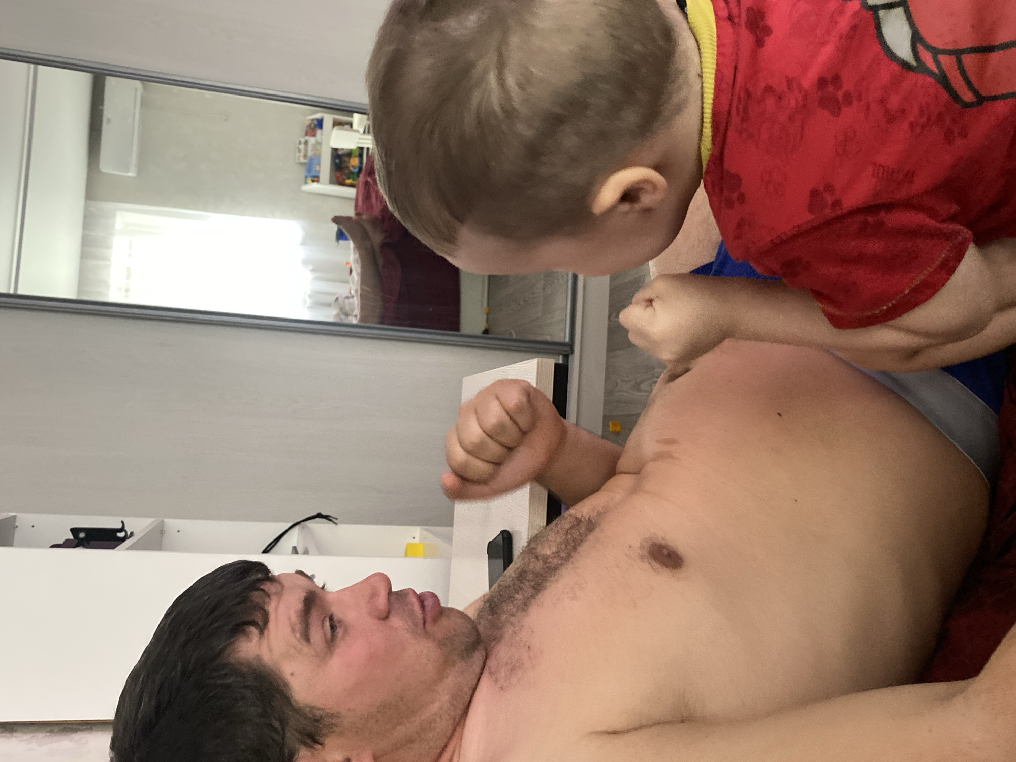 The image captures a playful moment between an adult male and a young child in an indoor setting. The adult male is lying on his back, partially shirtless, with dark hair and some chest hair. He is looking upwards and slightly to his right, towards the child, with an engaged expression. His right arm is bent, and his fist is clenched, resting near his shoulder. He appears to be wearing grey underwear, visible at his waist.
Leaning over the man, head down, is a young child with light brown hair. The child is wearing a red t-shirt with a yellow trim around the neck/shoulder, adorned with a subtle pattern of repeating motifs, possibly including stylized letters, numbers, or cartoon elements like paw prints. The child's hands are also clenched into fists, positioned near the man's midsection, suggesting a playful wrestling or boxing interaction.
The background shows a brightly lit room. To the left of the man, a white cabinet or shelf unit is visible, holding a black rectangular object (possibly a phone or remote control) and other small items. Above this, a large rectangular mirror reflects parts of the room. In the mirror's reflection, a white air conditioning unit is mounted on a light-colored wall, and a bright window with horizontal blinds is visible, indicating it is daytime. Further back in the reflection, what appears to be a shelf unit with various colorful objects, possibly toys or books, can be seen. The man is lying on a surface covered with red fabric, likely a bed or sofa. The overall scene conveys a warm, affectionate, and playful interaction between the two individuals.
