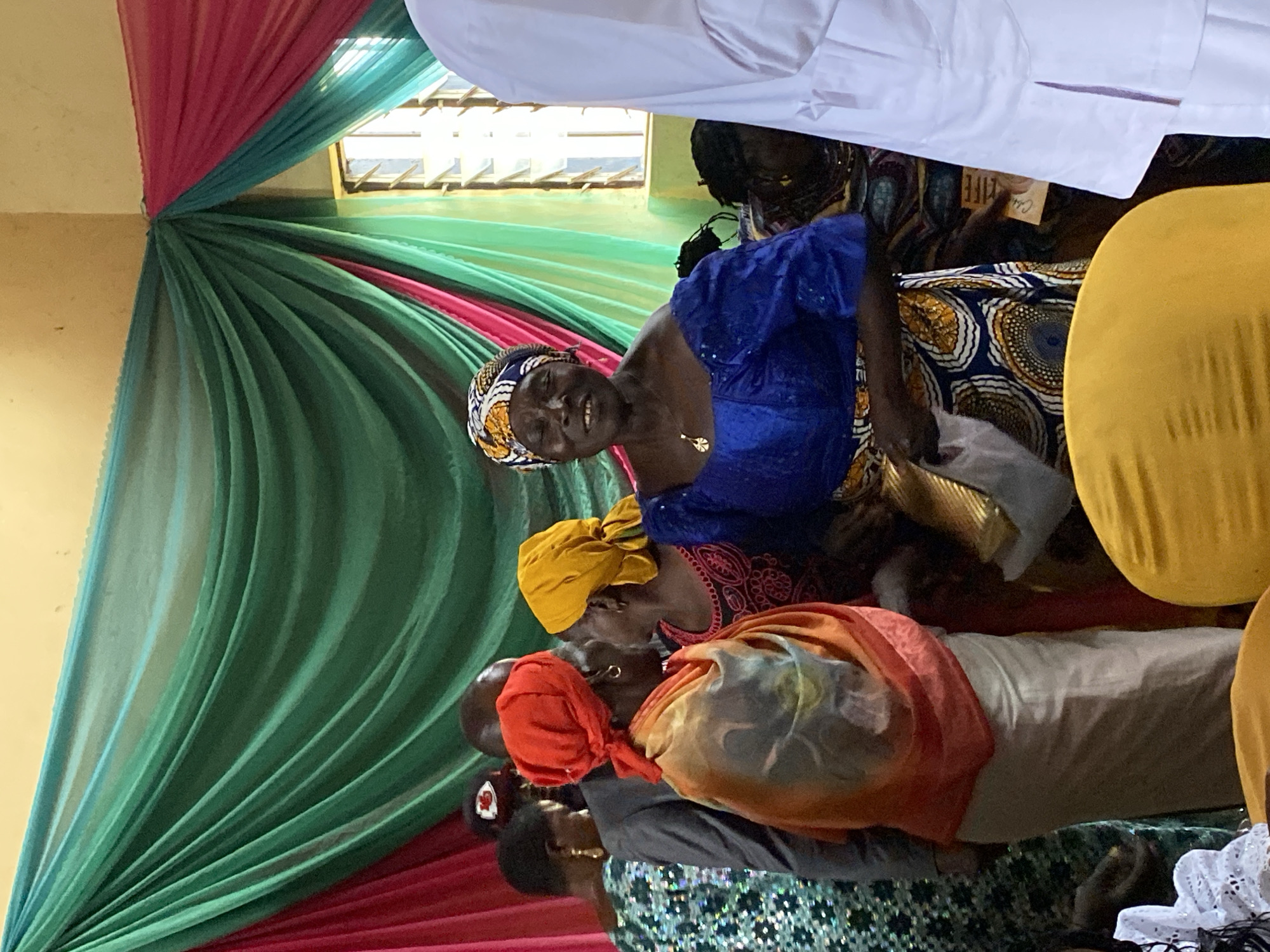 The image depicts an indoor gathering, possibly a celebration, with a backdrop of draped pink and green fabric. Several individuals are present, dressed in vibrant, traditional African attire. In the foreground, a woman in a blue off-the-shoulder top and a patterned skirt is smiling and looking towards the camera. To her left, another woman wears a yellow headscarf and is engaged in conversation with the woman in blue. Partially visible behind them are other attendees, including a person in a grey suit and a red headscarf. The environment suggests a community event, likely in Riyom, Nigeria, given the cultural context and the presence of African fabrics and clothing.