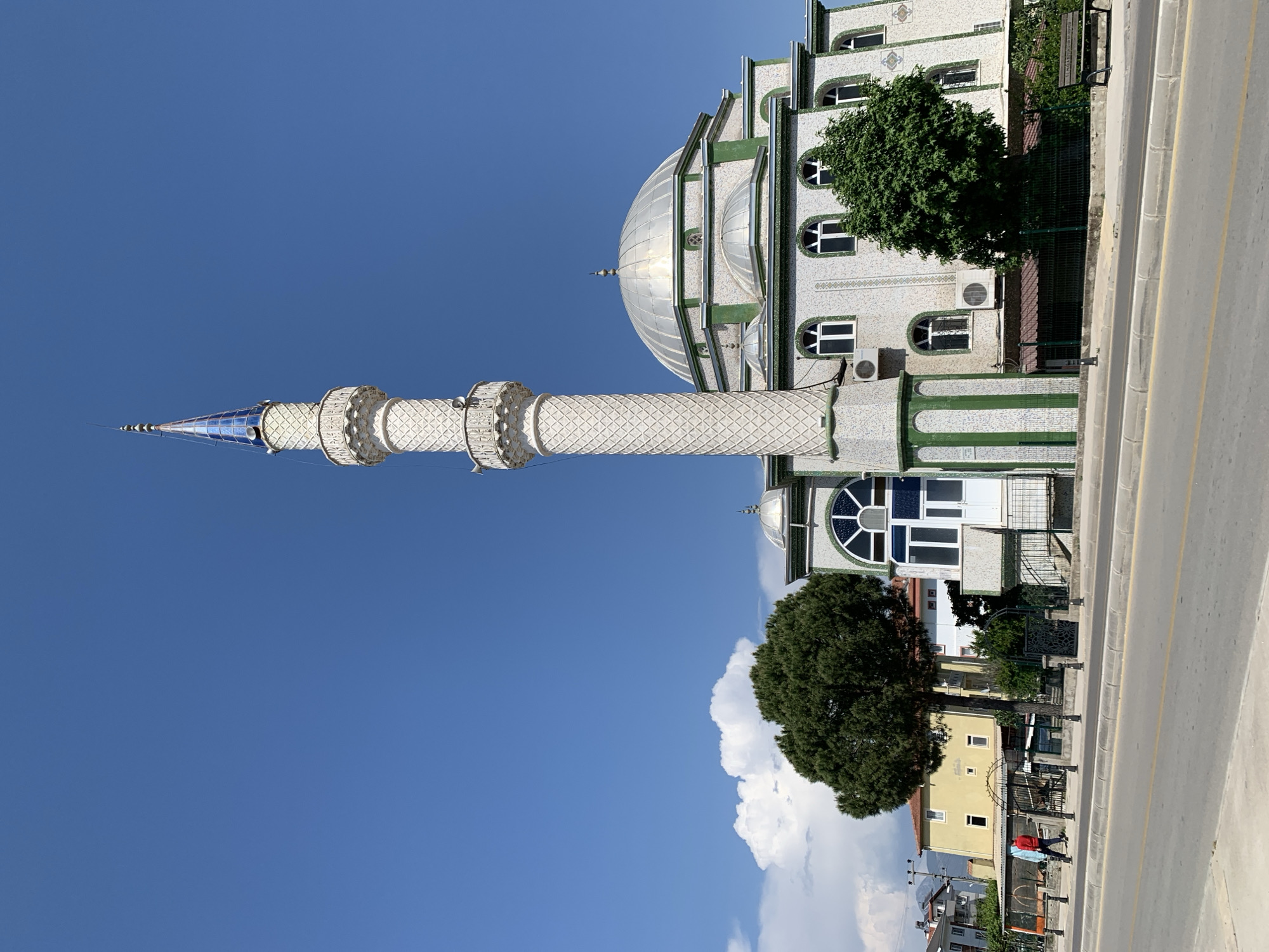 This media file captures a bright, sunny daytime scene featuring a mosque and its surroundings. The image, which appears to be rotated 90 degrees clockwise, shows a prominent mosque with a minaret, set against a clear blue sky.
The mosque building is predominantly white with striking green architectural accents and trim, giving it a tiled or mosaic appearance. It features numerous arched windows with white frames. A large, shimmering silver dome tops the main structure, accompanied by smaller domes visible behind it. Several air conditioning units are mounted on the exterior walls of the mosque.
To the left of the main mosque building stands a tall, slender minaret. It is also white with intricate patterns and has two circular balconies near its top. The minaret culminates in a pointed spire that is blue and silver, reflecting the sunlight.
In front of the mosque, a wide, grey road with yellow lane markings runs parallel to the building. A sidewalk borders the road. Several trees with lush dark green foliage are visible; a large, full tree stands near the mosque, and another is closer to the foreground near a yellow building. Beyond the road, in the mid-ground, there are residential-style buildings, including a distinctive yellow one. A person, partially visible and wearing a red shirt, can be seen near these buildings.
The weather is bright and sunny, indicated by the clear, deep blue sky with a few fluffy white cumulus clouds. There is no discernible text visible in the image. The architectural style of the mosque suggests a location in a country with Islamic heritage, consistent with Fethiye, Turkey.