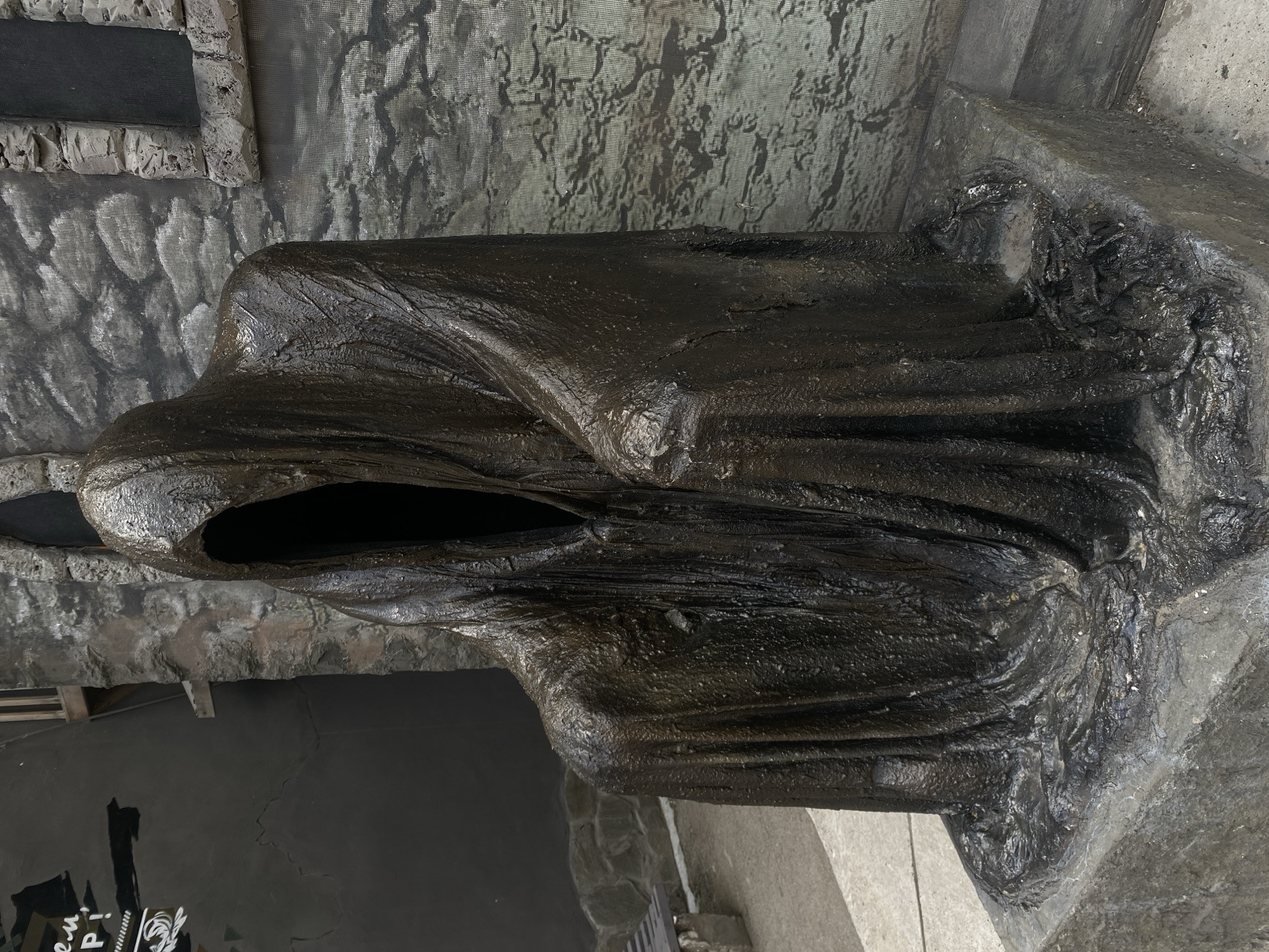 A life-sized, dark, textured statue of a robed, hooded figure sits on a stone base. The figure's hood obscures its face, creating a deep shadow where the face would be. The draped robe is detailed with folds and a rough, almost wet-looking texture. The statue is positioned outdoors, in what appears to be an urban environment. To the left of the statue, a dark wall with some white graffiti and a wooden beam is visible. Behind the statue, a grey, textured wall mimics stonework, with a rectangular opening that resembles a window frame. A paved walkway and a concrete bench are also present in the foreground and to the left. This scene is located in the city of Perm, Russia.