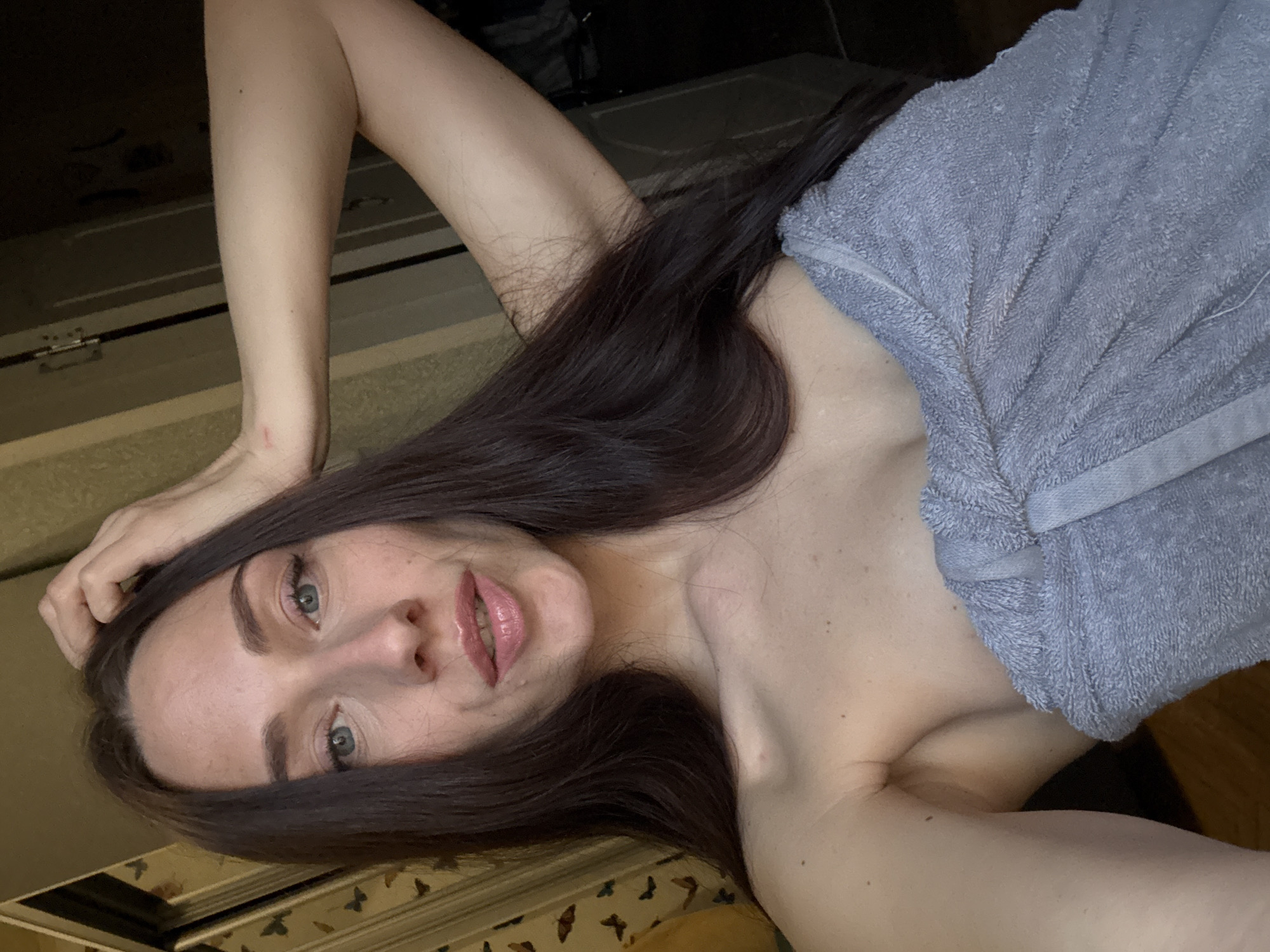 A woman with long, dark hair and light eyes is lying down, partially covered by a gray towel. She is looking at the camera with a slight smile. Her right hand is touching her hair. The background shows a decorated surface with butterfly patterns and wooden flooring. The location is not possible to determine from the image.