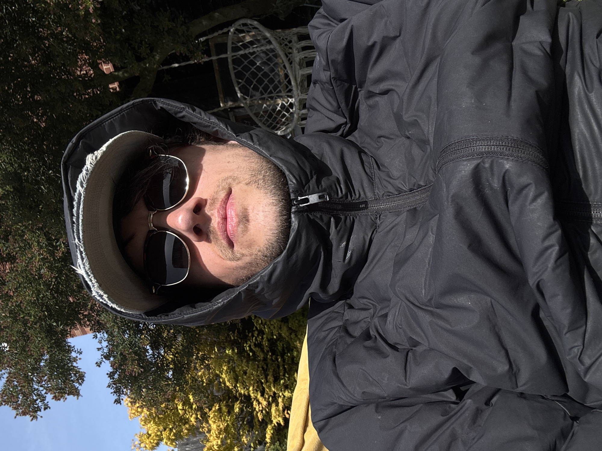 A medium close-up, rotated 90 degrees, shows a person outdoors in South Kesteven, United Kingdom. The person wears a black hooded down jacket with a visible zipper near the neck. They also wear round-framed sunglasses and a hat with a light-colored brim. The person's face is visible, including their nose, lips, and stubble. Behind the person are trees with green and yellow leaves against a blue sky. A white metal structure is also partially visible in the background. The lighting is bright and sunny.