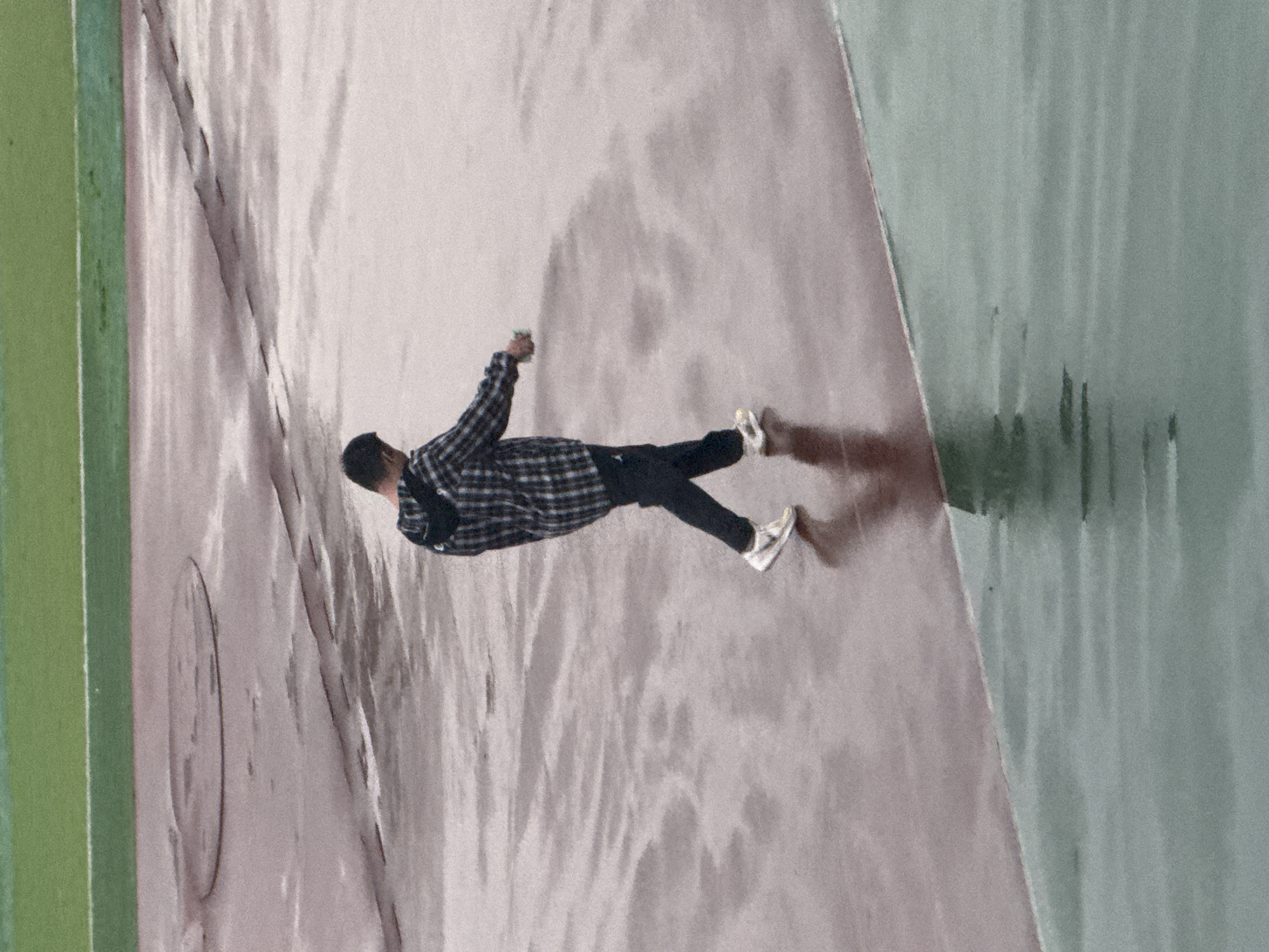 A photo taken outdoors in Zhongtun, China, presents a single individual, a male with black hair, captured from an elevated perspective. He is positioned on what appears to be a public thoroughfare or open space, clad in black pants, white sneakers, and a black and white checkered, long-sleeved shirt. He holds an item in his extended left arm, which is raised away from his body. He casts a shadow toward the right side of the frame. The surface he stands on is a light, grayish-purple color. To the left of the man is a vertical, green beam. To the right of the man is a vertical, grey-green area.