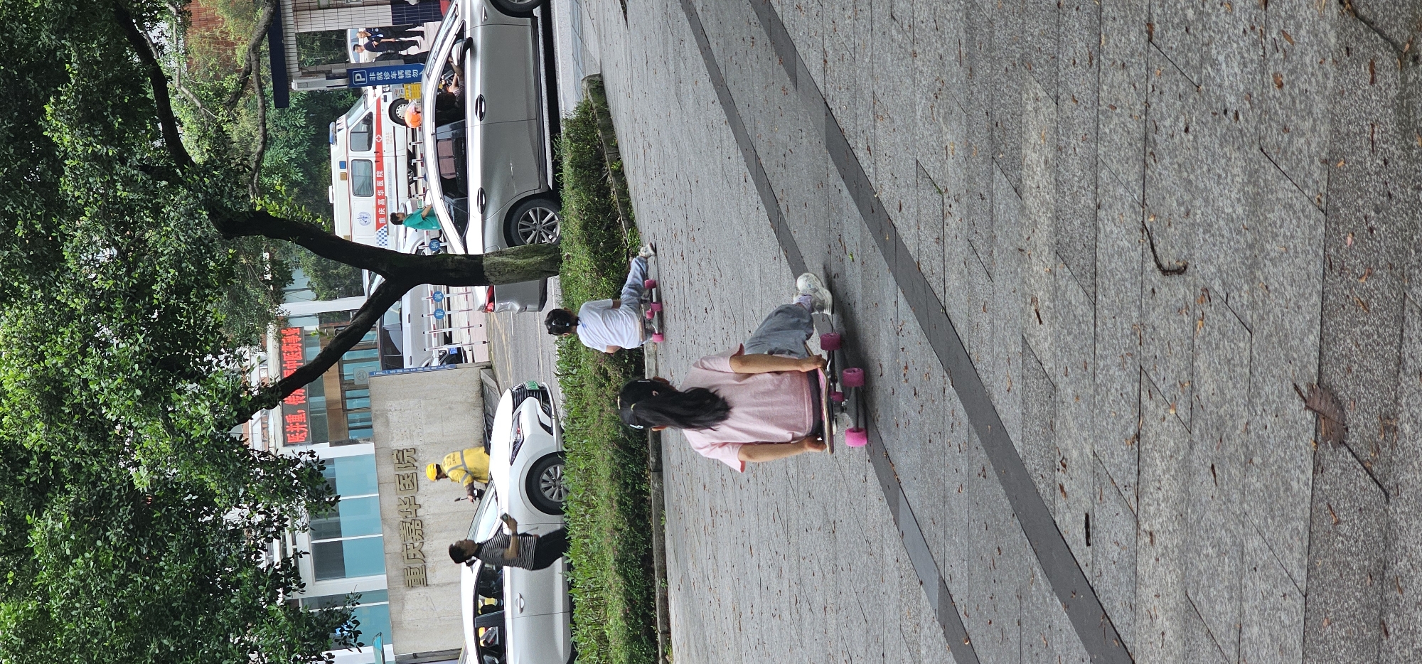 Here's a description of the image:
The image shows two people seated on skateboards, positioned on a paved sidewalk near a street. The first individual wears a pink top, while the second person is dressed in white. A low hedge separates the sidewalk from the street, where vehicles are parked, including an ambulance.
In the background, a building is visible with text in Chinese; specifically, "重庆嘉华医院 (Chongqing Jiahua Hospital)" indicating the location of the Jiulongpo District, Chongqing, China. A tree with dense foliage is present on the left side of the frame.