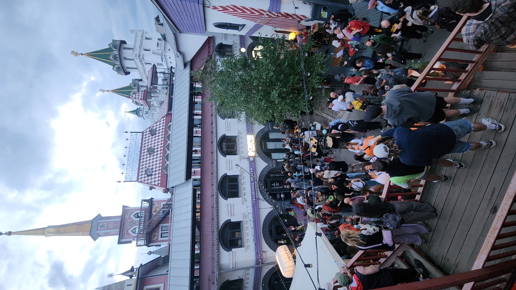 The image depicts a crowded outdoor venue in what appears to be the Izmailovo Kremlin in Moscow, Russia. The unique architecture of the Kremlin, with its colorful, fairytale-like towers and decorative facades, is visible. A large crowd of people is gathered in the courtyard below. The crowd appears to be an audience at an outdoor event. Many people are standing and moving around. A tree is in the center of the courtyard. A building with multiple windows and signs is near the crowd. The photograph is taken from a slightly elevated perspective, possibly from a balcony or a raised platform with wooden planks and railings. The sky above is overcast.