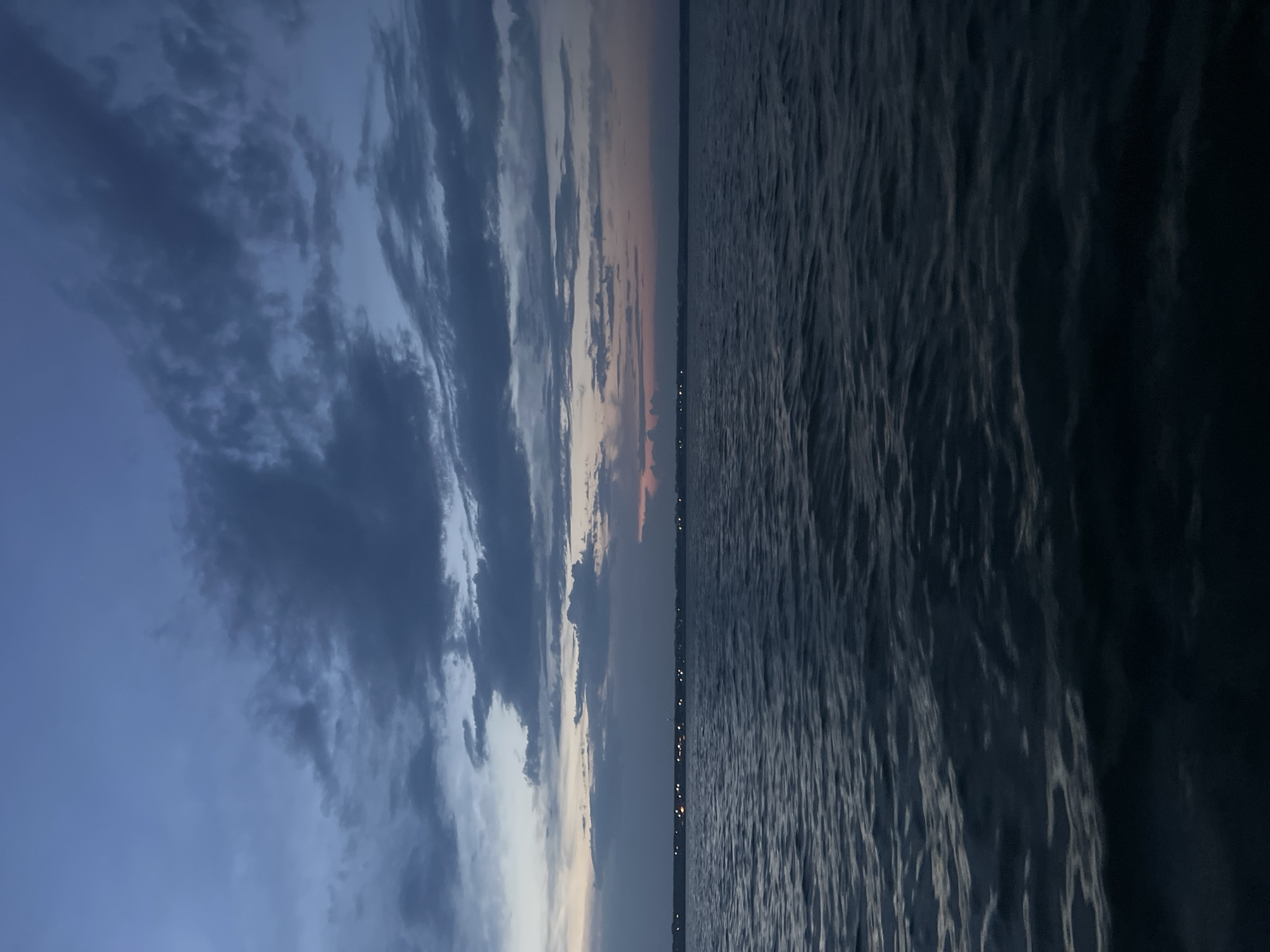 The image depicts a twilight scene over a body of water, likely a lake or a wide river, with a distant shoreline visible. The sky is a gradient of deep blues and grays, with scattered, textured clouds that suggest the transition from day to night. A faint band of pink and orange hues is visible near the horizon, hinting at the last remnants of sunset.
Along the distant shoreline, a line of small, bright lights is visible, suggesting a settlement or a town. The water in the foreground is dark and choppy, with ripples and small waves reflecting the dim light of the sky. The overall mood of the image is serene and tranquil, evoking a sense of calm as day gives way to night.
The location in the city of Novopichugovo, Russia, is not discernible from the visual information provided in the image. There are no identifiable landmarks, signs, or specific geographical features that would allow for its precise identification within Novopichugovo. The scene is generic enough to be found in many locations with similar geographical characteristics. There are no people or discernible actions taking place in the image.