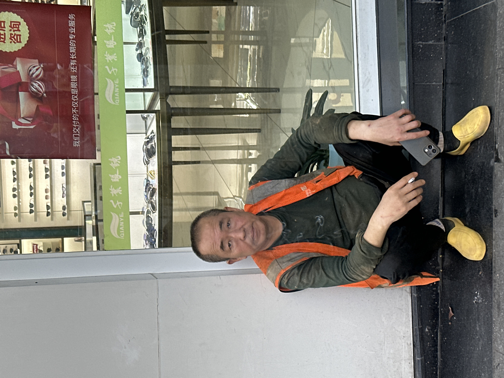A man is depicted sitting on the ground, leaning against a white wall or panel, outside a shop in Jiangjin, China. He is wearing a dark green long-sleeved shirt, an orange and grey safety vest, black trousers, and distinctive bright yellow shoes. Black gloves cover his hands, with visible dirt on them and scratches on his left forearm. He holds a dark-colored smartphone in his left hand and a lit cigarette between the fingers of his right hand. His expression is neutral to slightly weary, as he looks towards the viewer.
Behind the man is a large glass storefront, showcasing numerous eyeglasses on display shelves inside. Reflections are visible on the glass surface. To the left of the man, integrated into the storefront design, are two prominent signs. One is a red sign featuring a festive gift box graphic, with Chinese text that reads "咨询" (Consultation) and a longer slogan: "我们交付的不仅仅是眼镜还有长期的专业服务" (What we deliver is not just glasses, but also long-term professional service). Below this, a green horizontal strip sign displays "QIANYE" and "千葉眼鏡" (Qianye Eyewear), which is repeated further along the sign. The man is resting on a dark, possibly granite or tiled, floor, with a white wall behind his upper body. The lighting suggests it is daytime, with diffused light.