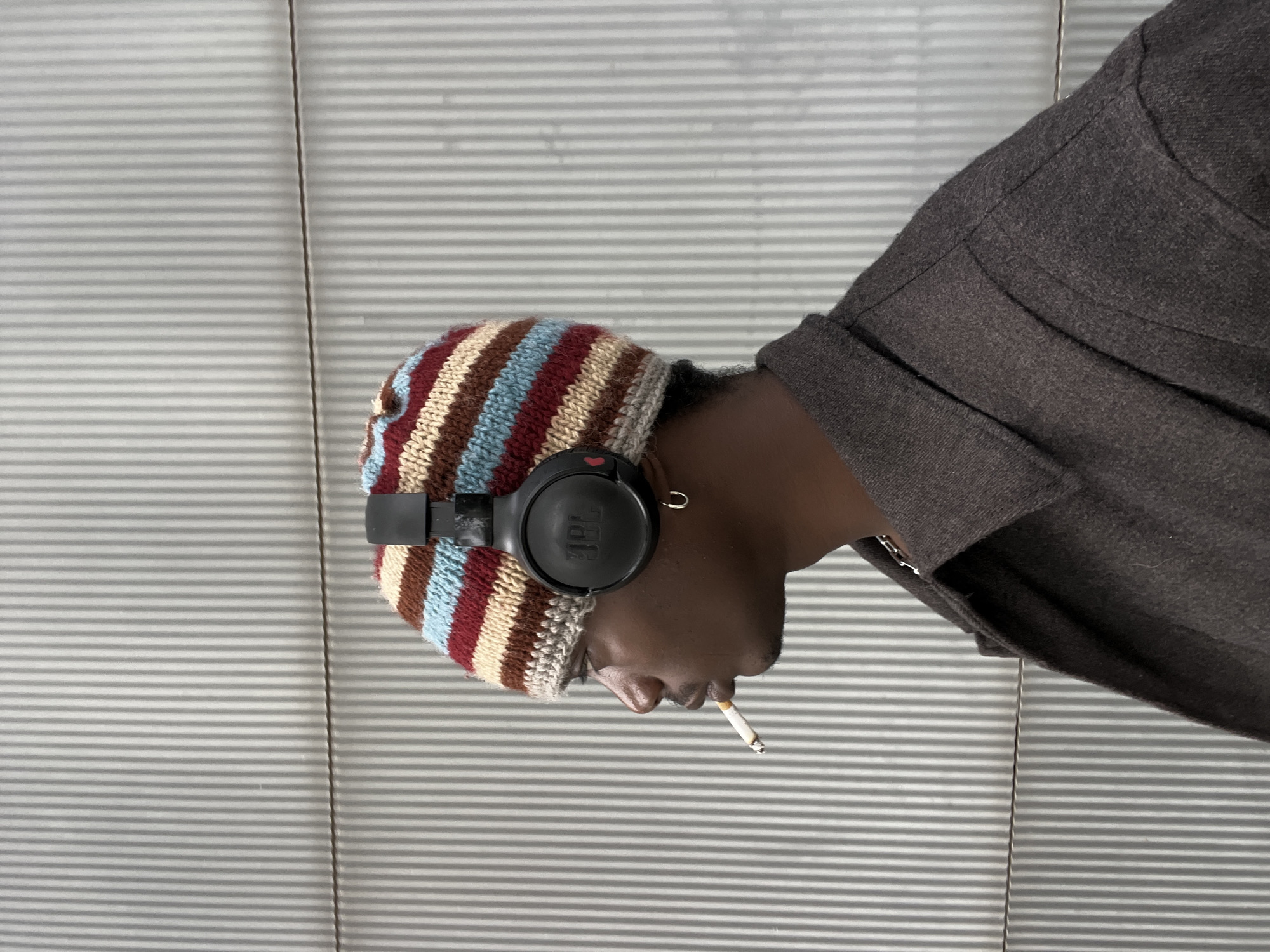 The image presents a rotated profile view of a person, possibly male, with dark skin, captured from the head down to the upper torso. The individual is wearing a distinctive knitted beanie with horizontal stripes in shades of maroon, light blue, brown, and beige/cream. Black over-ear headphones, clearly branded with the "JBL" logo on the earcup, are worn over the beanie. A small golden hoop earring is visible on the left ear.
The person is dressed in a dark-colored jacket or coat, which appears to be a deep brown or dark grey. A lit cigarette is held between their lips, with a visible ember and a wisp of smoke, indicating they are actively smoking. Their gaze is directed downwards and slightly away, suggesting a moment of contemplation or relaxation.
The background features a uniform surface with numerous thin, horizontal grey lines, resembling closed venetian blinds or a textured wall paneling. A faint vertical line, possibly a seam or a thin cable, dissects the background. The lighting is soft and diffused, offering no specific cues regarding the time of day or outdoor weather. While the scene lacks overt architectural or environmental markers, the context provided situates this moment in Barcelona, Spain. The image conveys a personal moment of leisure, combining music listening with smoking.