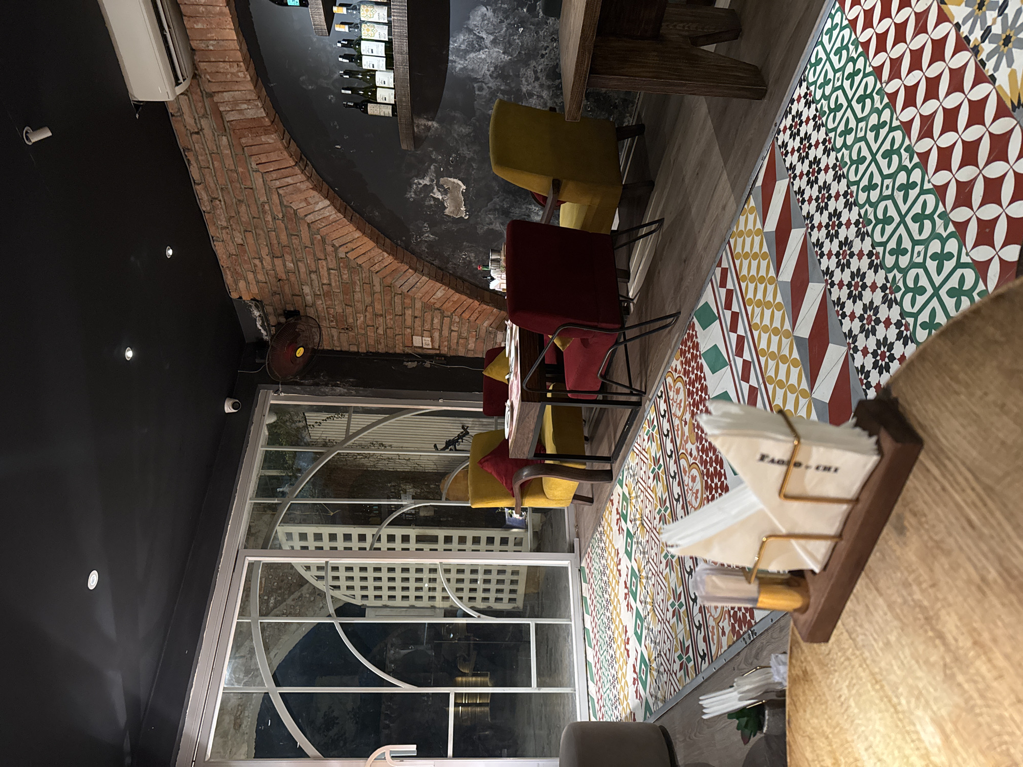 This image shows the interior of a dimly lit restaurant in Hanoi, Vietnam. The scene features a mix of rustic and modern elements, with exposed brick walls and a dark ceiling contrasting with colorful patterned floor tiles and sleek furniture.
In the foreground, a wooden table holds a napkin dispenser with the visible text "FABIO CHI." To the right, a wooden counter with a reflective surface is partially visible.
Further back, several red and yellow chairs are arranged around tables. Some chairs have red cushions. A large window with a white lattice design looks out onto what appears to be an outdoor area, though it is difficult to discern specific details. An air conditioning unit is mounted on the dark wall above the brickwork. A small, circular security camera is visible on the upper left wall. A red fan is positioned on the floor near the brick wall.
The lighting suggests it is likely evening or nighttime. The overall ambiance seems relaxed and somewhat upscale. No people are clearly visible in the frame.
