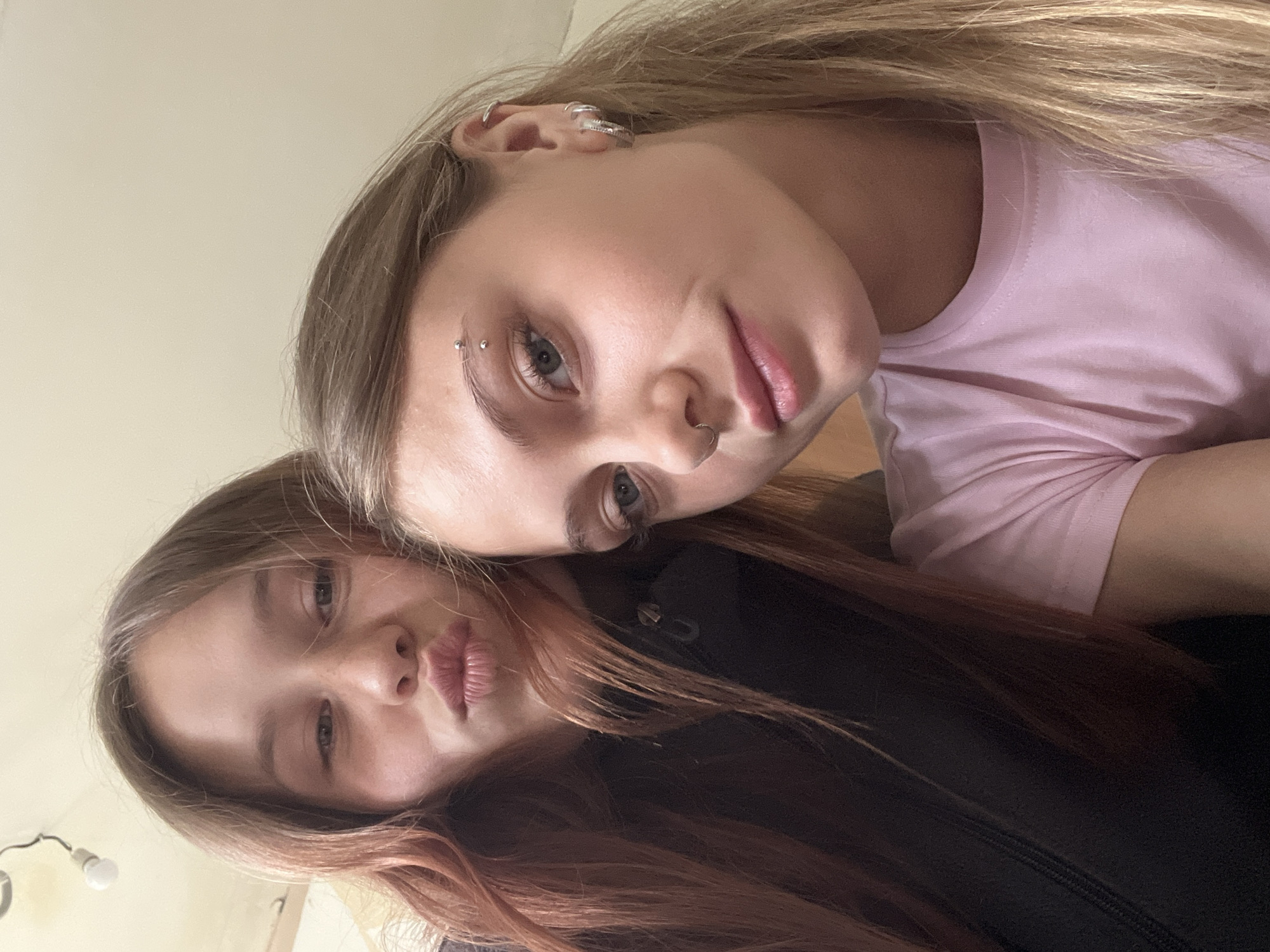 Two young women pose closely together for a selfie. The woman on top has blonde hair, light skin, and is wearing a pink shirt. She has a pierced eyebrow, nose ring, and multiple earrings. The woman below has brown hair with reddish highlights and is wearing a black jacket. The background is out of focus, but appears to be an indoor space with light-colored walls. There is a light fixture visible in the lower left corner. There is no evidence to suggest that this image was taken in Фоки, Russia.