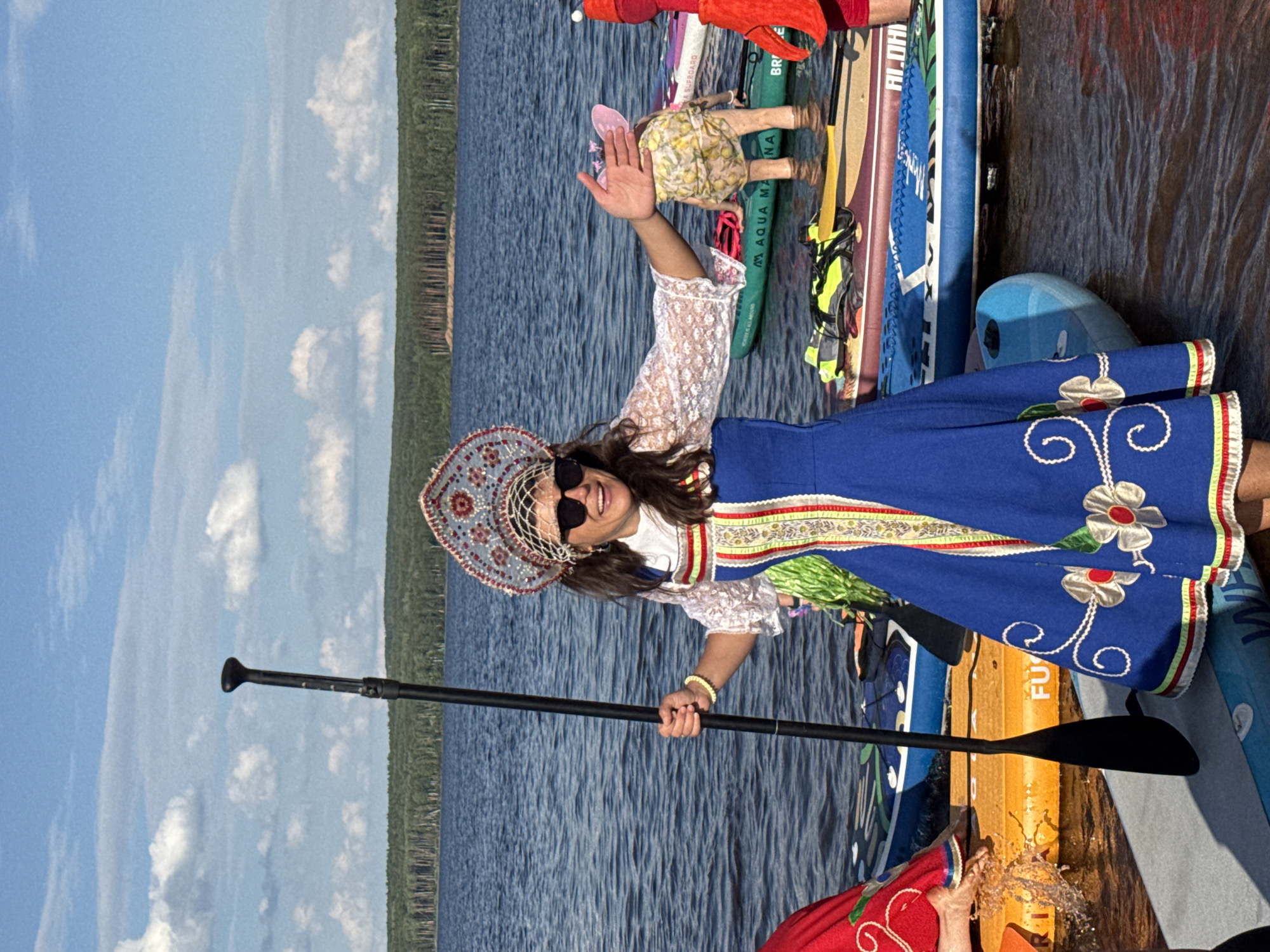 A woman in traditional Russian attire, including a kokoshnik headdress and a blue dress with embroidered floral patterns, stands on a paddleboard in a body of water. She is holding a paddle and smiling at the camera, with her right hand extended. Behind her, other paddleboards are visible, some with people on them. One person is wearing a yellow and white lemon-patterned outfit and has pink fairy wings. Another person on the right is wearing a red costume with a hood. The water appears calm, with ripples on the surface. In the background, a dense forest lines the shore under a partly cloudy blue sky. The scene is set on the Kama River in Chaikovsky, Russia, a city known for its water sports and scenic natural beauty.