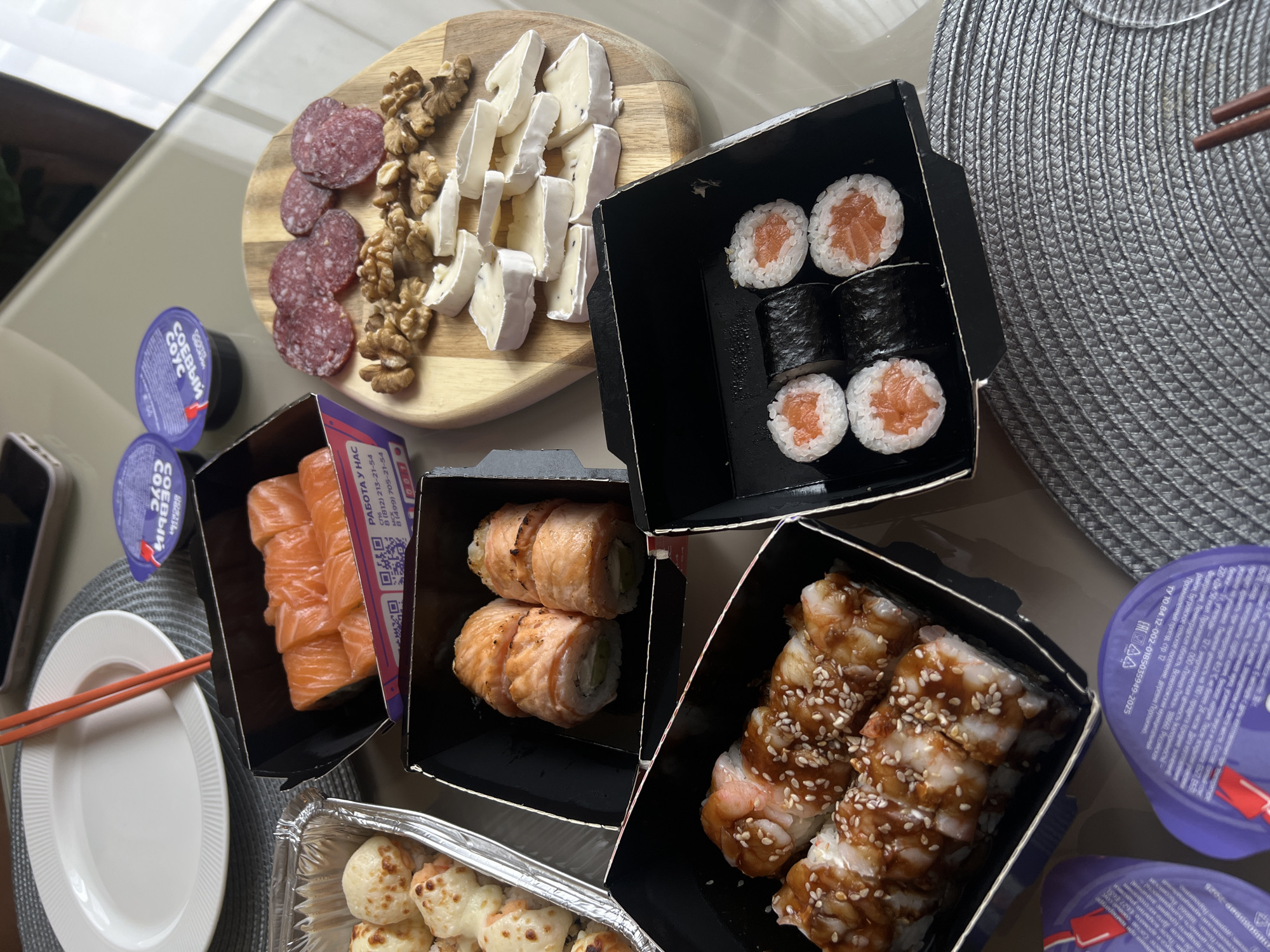 The image shows a table laden with a variety of food items, including sushi, a cheese and charcuterie board, and small tubs of what appears to be soy sauce. There are several containers of sushi, with different types including maki rolls, nigiri, and possibly baked rolls. The cheese board features brie, walnuts, and salami. Chopsticks and a white plate are set on a circular woven placemat. A smartphone is partially visible on the table. A label is present on one of the sushi containers with Russian text referencing a job opportunity, phone numbers (including a 812 area code), and the company "Full Shelf" located in the Leningrad Oblast, near St. Petersburg, Russia (not Khimki). The label includes a manufacturing date.