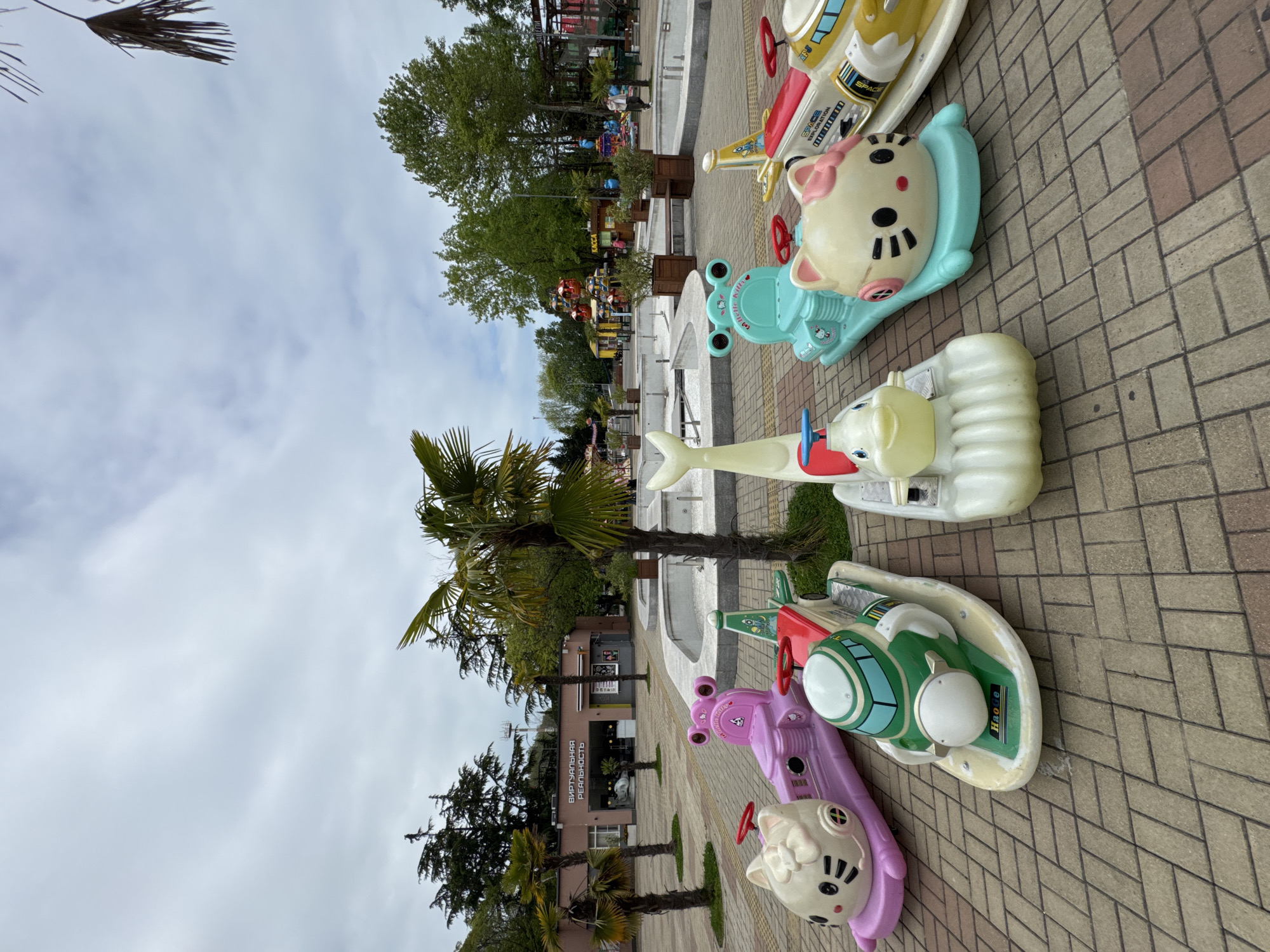The image shows a row of kiddie rides on a brick-paved promenade in Sochi, Russia. The rides are various animal and vehicle shapes in bright colors. A palm tree and other trees are visible in the background. In the distance, there is a small fairground. The sky is overcast. A building with a sign in Russian can be seen along the path.