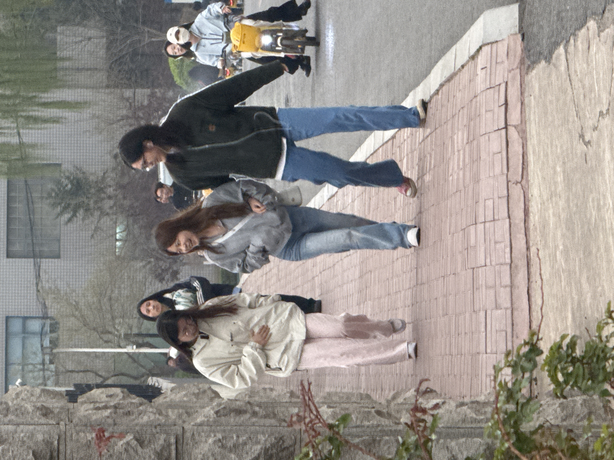 The image depicts an outdoor street scene in Zhongtun, China, likely during an overcast or hazy daytime. The perspective is slightly elevated, looking along a paved sidewalk that runs parallel to an asphalt road.
In the foreground, several people are walking on the reddish-brown brick sidewalk. Closest to the viewer, a person stands wearing a light cream-colored jacket and light pink pants. Behind them, another person in a grey hooded sweatshirt and blue jeans walks with a light-colored shoulder bag, looking towards the camera with a subtle expression. Further back on the sidewalk, a third person is seen walking in a dark jacket and blue jeans.
On the road to the right, two individuals are riding a yellow scooter or small motorcycle. The driver wears a light-colored jacket and a white face mask, while the passenger also wears a light-colored jacket.
The sidewalk is bordered on the left by a low stone wall and on the right by the road. In the background, modern-style buildings with multiple windows are visible through sparse trees and shrubbery, suggesting an urban or campus environment. The lighting is diffused, indicating no direct sunlight and a generally calm atmosphere. There is no discernible text visible in the image.
