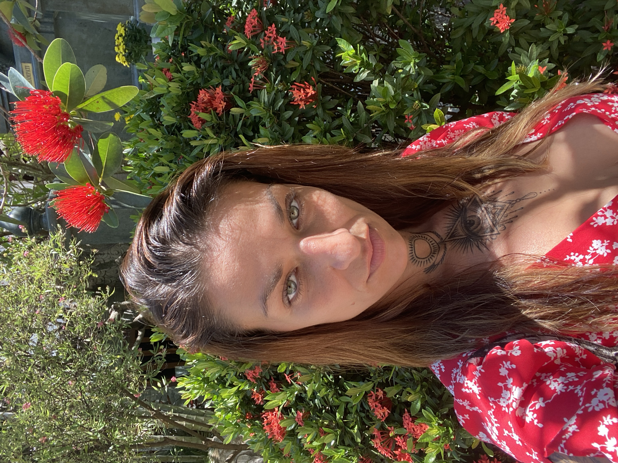 A woman with brown hair and green eyes is posing for a photograph in a garden. She is wearing a red and white floral dress. She has a tattoo on her neck and chest. Behind her are green bushes with small red flowers. To the left are green bushes with red, and yellow flowers. The exact location in the city of Dĩ An, Vietnam cannot be determined without additional information.
