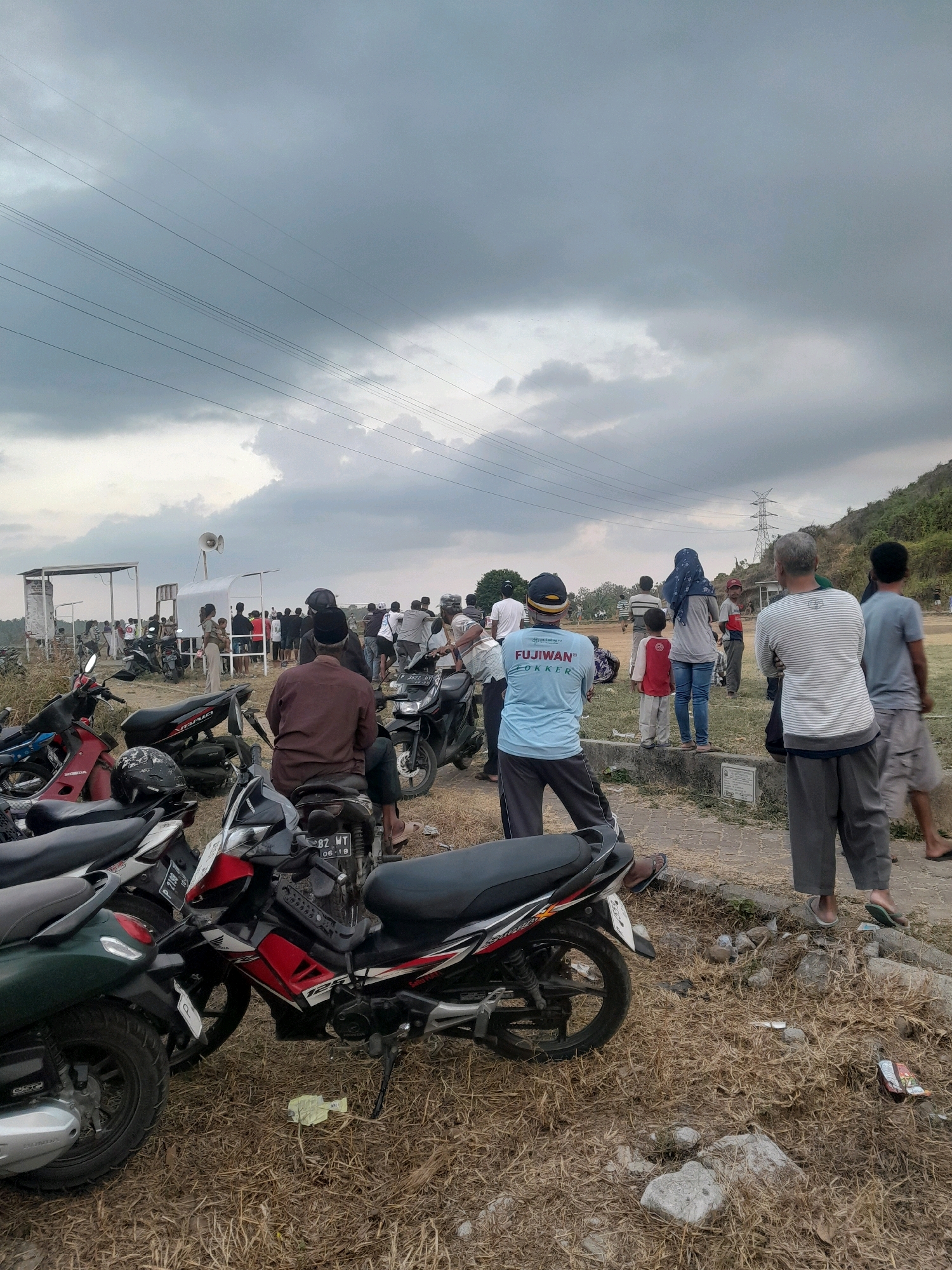 The image depicts a gathering of people in an outdoor, somewhat arid environment, likely in Genteng, Indonesia, given the context. The sky is overcast with dark clouds, suggesting an impending change in weather. A prominent feature in the foreground is a collection of parked motorcycles, including a red and black motorbike with the number plate "82 WT 06-19". In the background, a crowd of people are standing, some appearing to be watching an event or activity. There are structures that resemble temporary shelters or stalls, one of which has a loudspeaker mounted on top. Electrical power lines crisscross the sky, and a transmission tower is visible on a distant hill. The ground is covered with dry grass and scattered rocks. The overall scene suggests a communal gathering or event taking place in a rural or semi-urban setting.