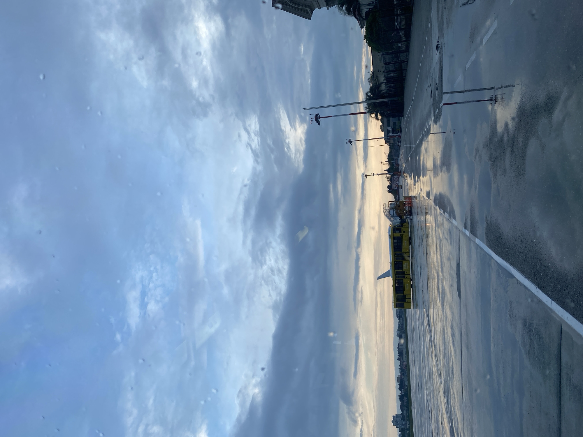 The image shows a view from inside a vehicle on a wet street in what appears to be Sochi, Russia. The sky is overcast with a mix of light and dark clouds. The street surface is reflective from the rain, and there are white lane markings along the road. Along the roadside are street lights and buildings. A yellow vehicle is parked along the road. There are droplets on the window of the vehicle from which the photo was taken.