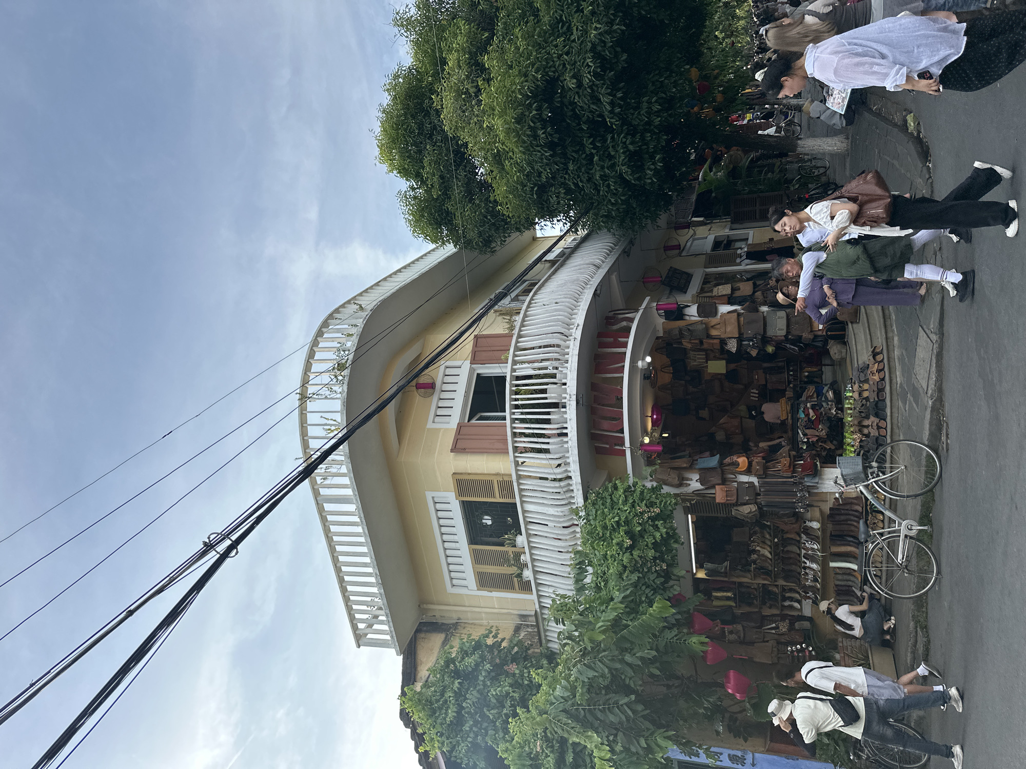 This image depicts a street scene in Hội An Ward, Vietnam, likely during the day with overcast skies. The foreground is dominated by a yellow building with a curved balcony railing and wooden shutters on its windows. Several large trees with dense green foliage flank the building and partially obscure its facade.
In front of the building, there's a shop displaying a large assortment of leather goods, including bags and shoes, arranged on shelves that extend out onto the street. A silver bicycle is parked near the shop entrance.
Several people are visible in the scene. In the upper right, two women are standing near the shop, one in a white shirt and the other in a darker outfit, seemingly looking at something in their hands. Further down the street, a person in a white hat and a white shirt with a black crossbody bag is walking, while another person in a white t-shirt and shorts is bent over, possibly interacting with the shop or something on the ground. Another person, wearing a white t-shirt and dark pants, is also visible near the shop. The overall atmosphere appears to be that of a busy commercial street with pedestrians browsing the shops.