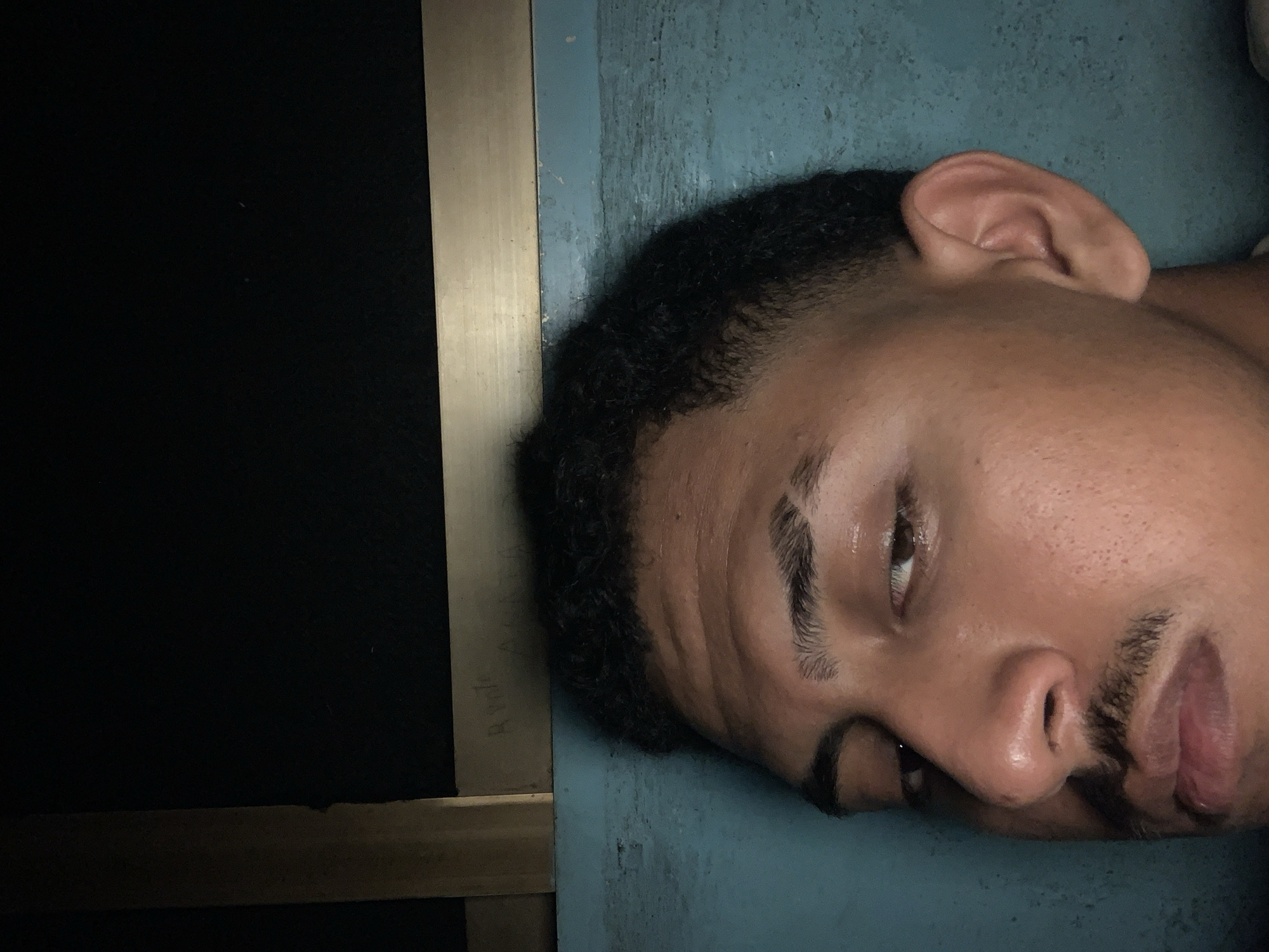 The image presents a close-up, portrait-style view of a young man, likely of mixed ethnicity, lying on his side. His head and the upper part of his shoulder are visible, resting against a textured, blue-green surface. He has short, dark, curly hair, well-defined eyebrows, a mustache, and a short beard or stubble. His right eye is partially open and looking slightly upward, while his left eye is mostly obscured. His mouth is slightly agape. A sheen is visible on his forehead and nose, which could indicate sweat, oil, or lighting conditions.
To the left of the man, a narrow, vertical strip, appearing to be light-colored wood or metal, separates him from a large, dark, almost black area that occupies the rest of the left side of the frame. This dark area suggests a deeply shadowed space or an enclosed structure. The lighting is dim, appearing to come from the top right, creating soft highlights on his skin and facial features.
The man's relaxed posture and partially open eyes suggest he is resting or possibly sleeping. There are no overt emotional expressions, but a sense of repose is conveyed. Faint, partially illegible text, which could be "R Ponto" or "R Ponte" followed by other characters like "A H", is barely discernible on the light-colored vertical strip next to his head.