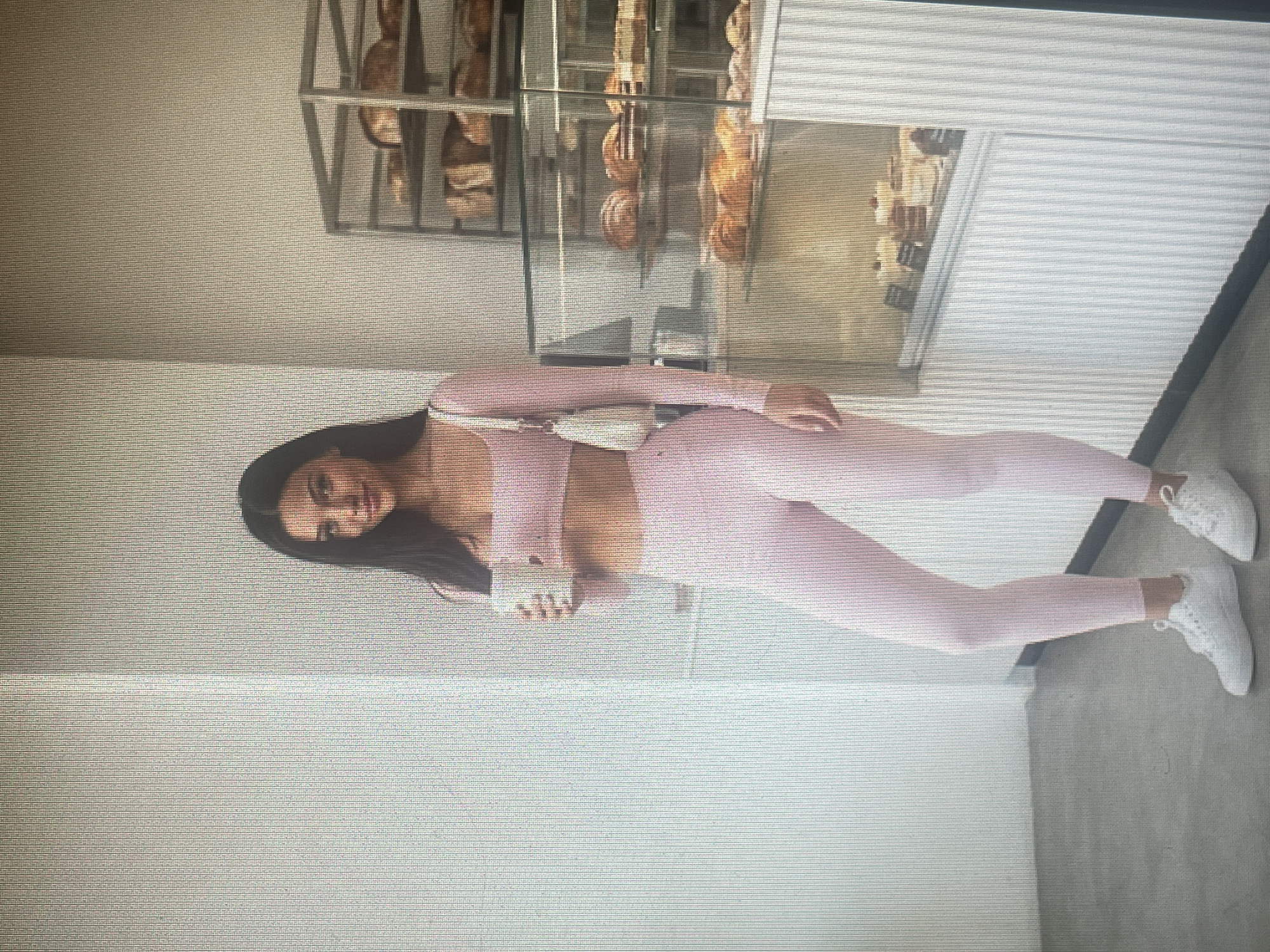 A young woman with long, dark hair stands in a bakery, holding a white mug. She is wearing a light pink two-piece athletic outfit consisting of a long-sleeved cropped top and leggings. A small, white purse is slung over her shoulder. She is wearing white sneakers. Behind her, shelves are stocked with baked goods, including bread loaves and pastries displayed in glass cases. The bakery has light-colored walls and a gray floor. The scene is well-lit, suggesting it is daytime. The specific location in Mantov, Czechia, is not discernible from the image.
