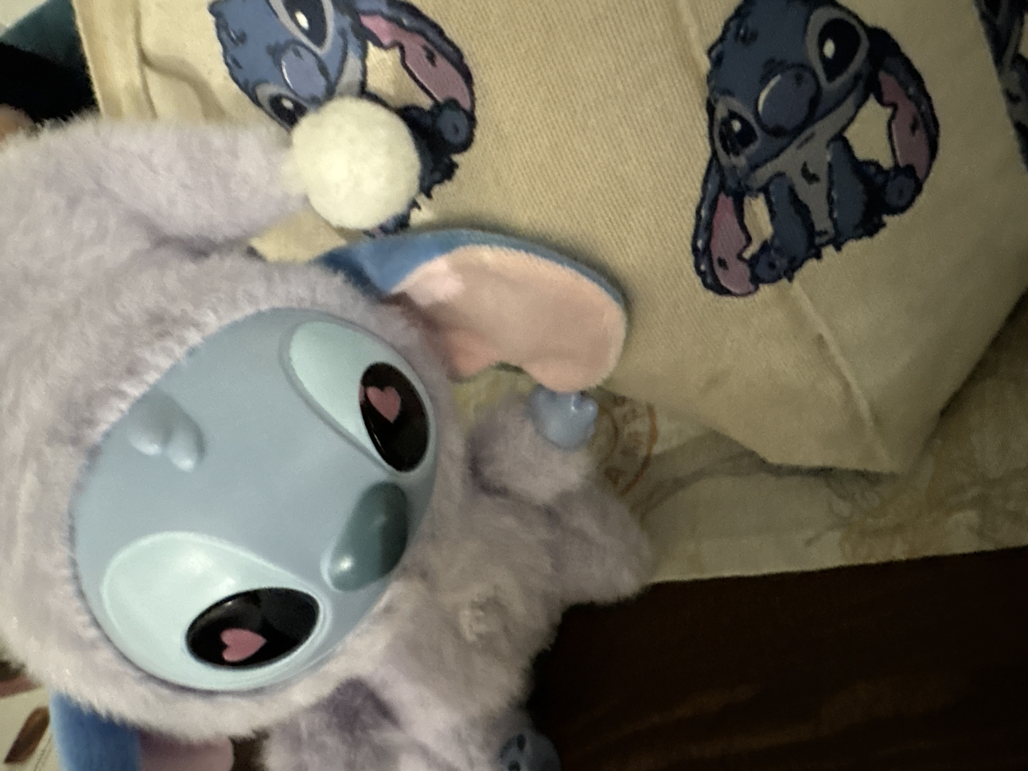 A plush Stitch toy dressed in a purple bunny costume is placed in front of a pillow with a printed image of Stitch. The toy has a blue plastic face with heart-shaped markings in its eyes. The environment appears to be indoors, possibly a home setting, with a darker surface visible in the lower right corner. The city of Unknown, Unknown is not discernable from the provided visual information.