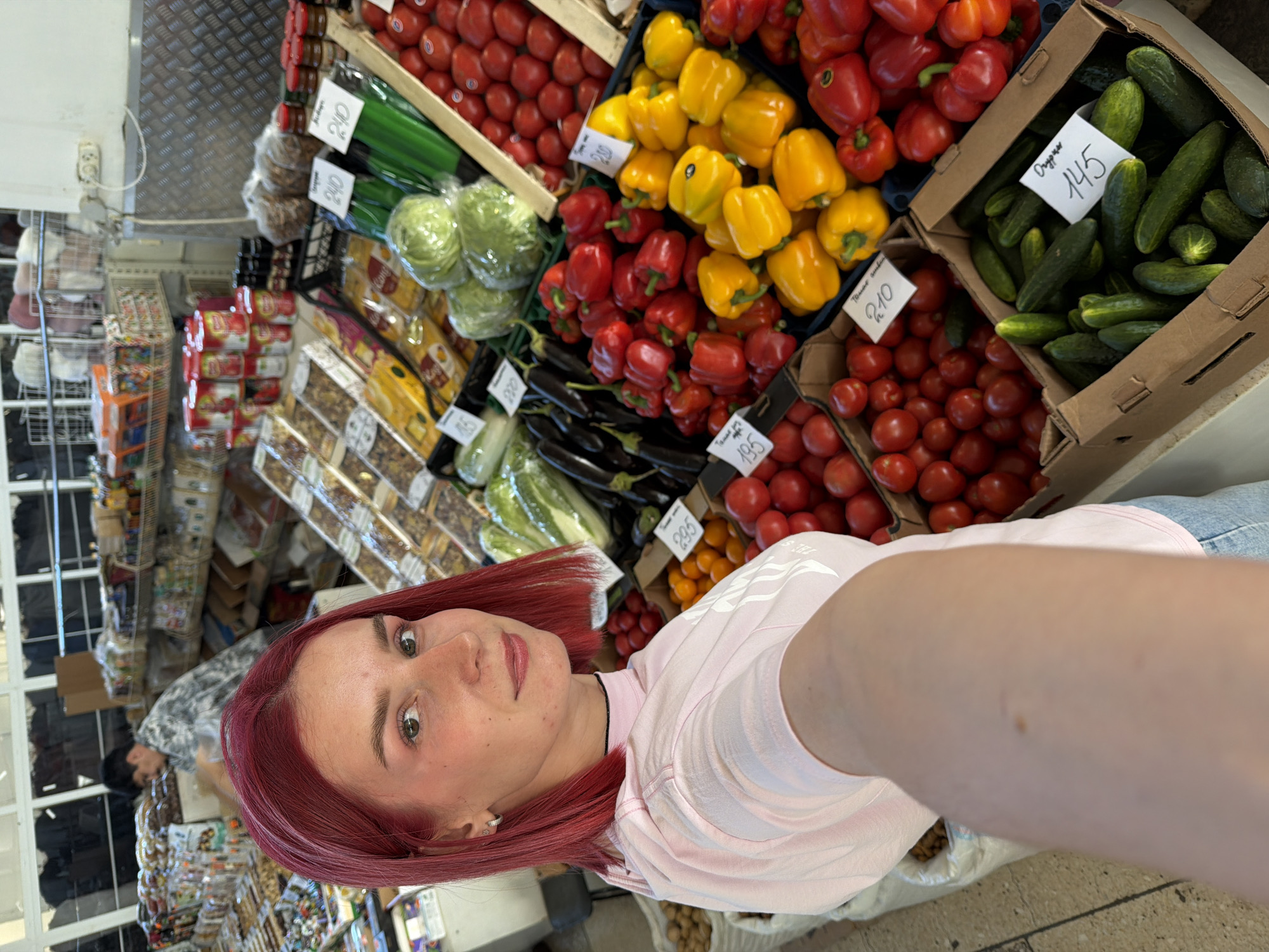 In Chaikovsky, Russia, a young woman with vibrant red hair takes a selfie at a marketplace. She is wearing a light pink hooded sweatshirt and blue jeans. Behind her, a man in a grey patterned shirt is visible, engaged in activity at a stall. The marketplace features numerous stalls laden with fresh produce, including tomatoes, bell peppers of various colors, cabbage, cucumbers, and eggplants. Packaged goods, canned items, and nuts are also displayed on shelves and in bins. The overall environment suggests a bustling open-air market or a well-stocked food market.