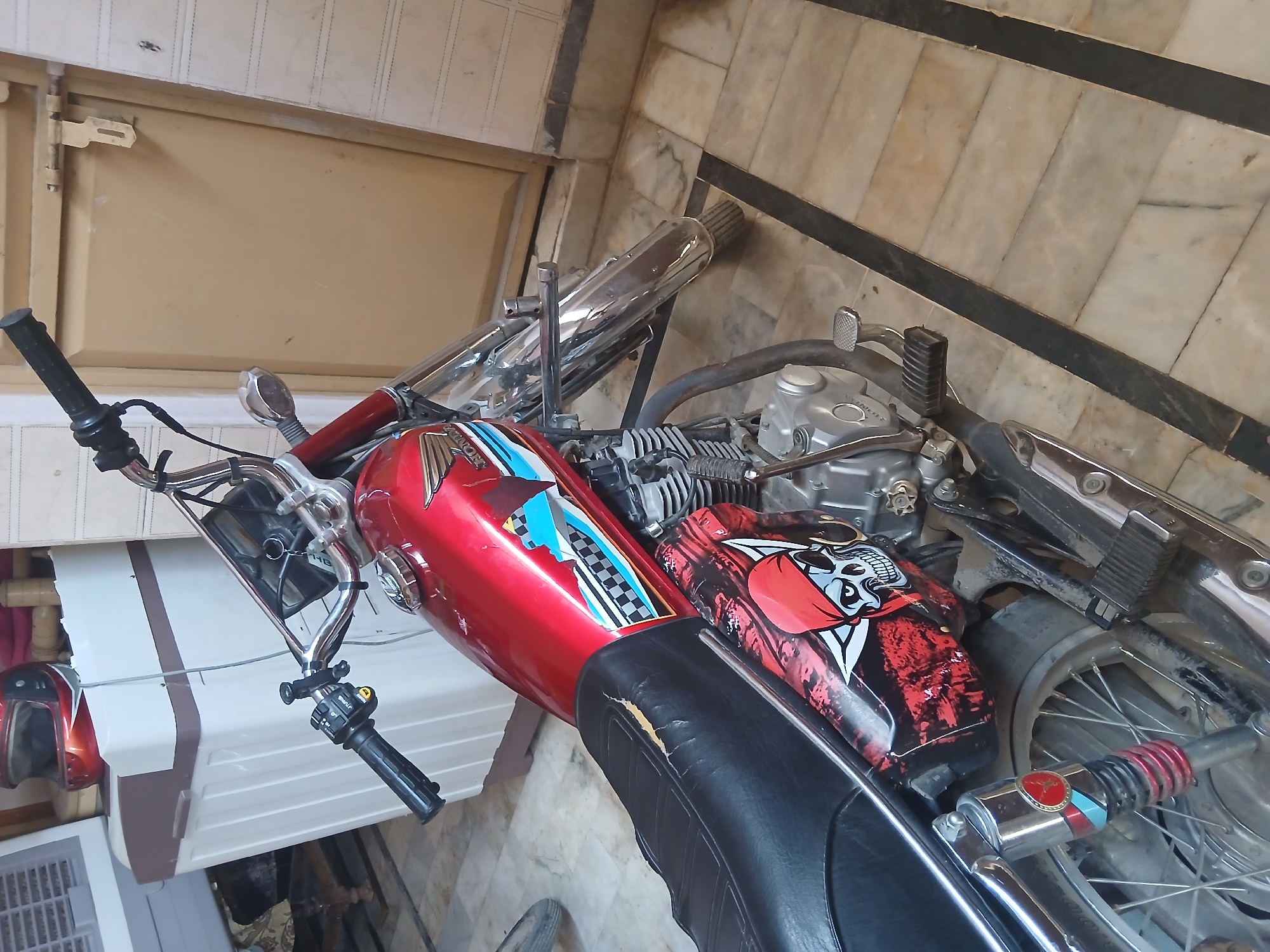 In Peshawar City Tehsil, Pakistan, a red motorcycle with a skull decal on its side is parked indoors on a tiled floor. The motorcycle has chrome accents, a black seat with visible wear, and is positioned next to a beige and brown washing machine. A motorcycle helmet, also red and black, rests on top of the washing machine. The background shows a beige door and a tiled wall.