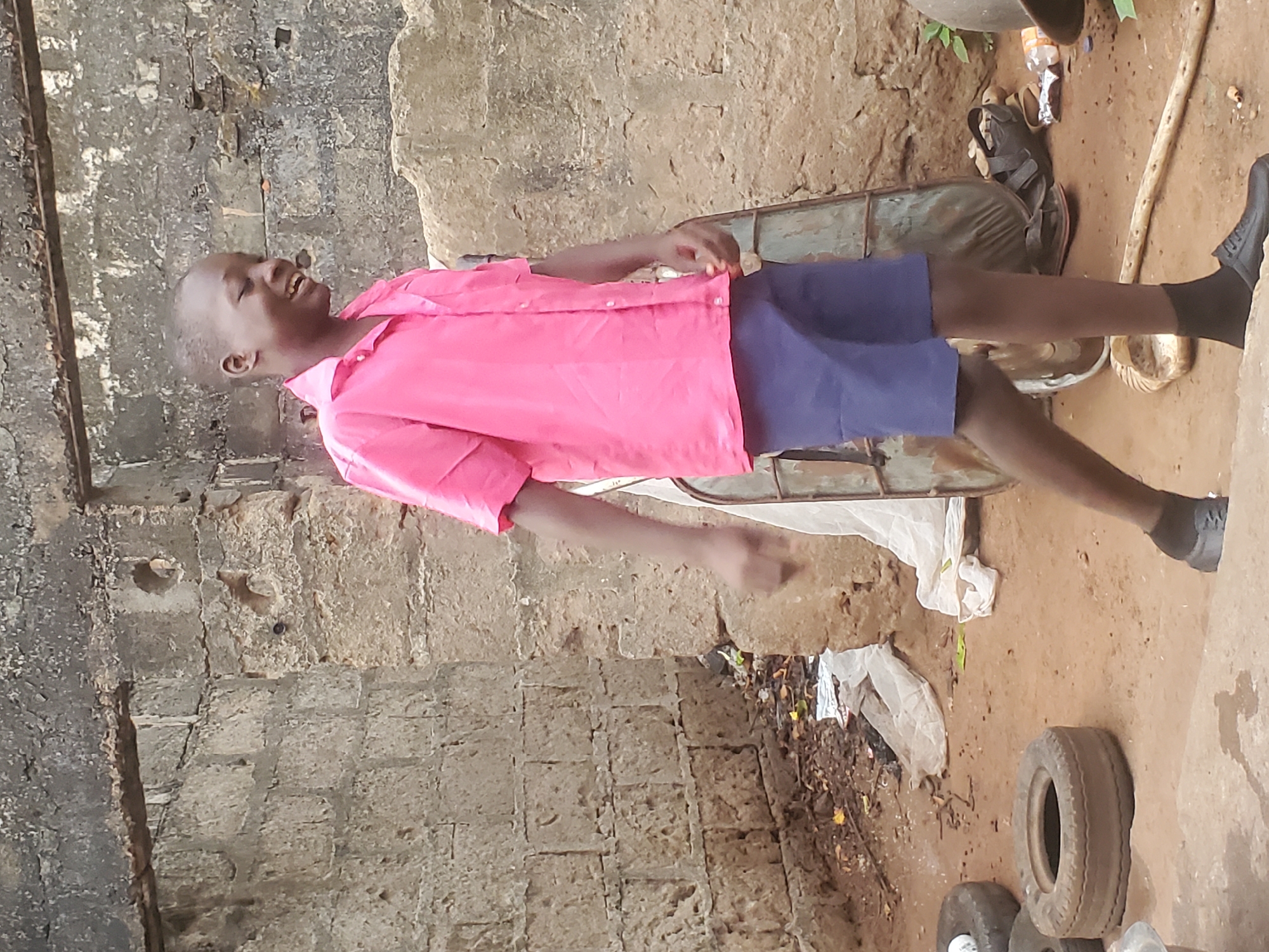 The image shows a young person standing outdoors in what appears to be a yard or compound. The person is wearing a pink shirt and dark shorts with black socks and shoes. The person is positioned towards the left of the frame with their head turned to the left and the body leaning to the right. The background includes a brick wall, and various objects like discarded tires, gardening tools and other debris scattered around. While the prompt provides the information the image depicts a location in Ukwu-Abwa, Nigeria, there is no additional information available from the image itself to support this assertion. The subject seems relaxed, leaning back.