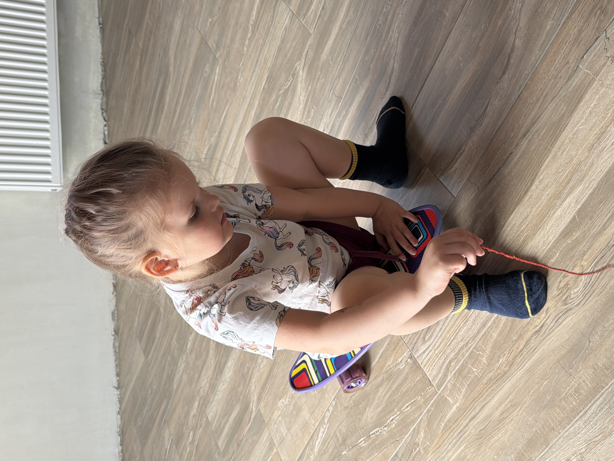 A child with light blonde hair is sitting cross-legged on a light brown tile floor, facing left. She is wearing a white t-shirt with a unicorn pattern and a maroon skirt. Her socks are dark blue with yellow stripes at the top. She is holding a red string and manipulating a small skateboard with a purple and multi-colored deck. The skateboard is under her legs. A white radiator is visible in the upper left corner of the frame. The location is in a residential building, but the city of Chaikovsky, Russia, cannot be determined from the provided information.