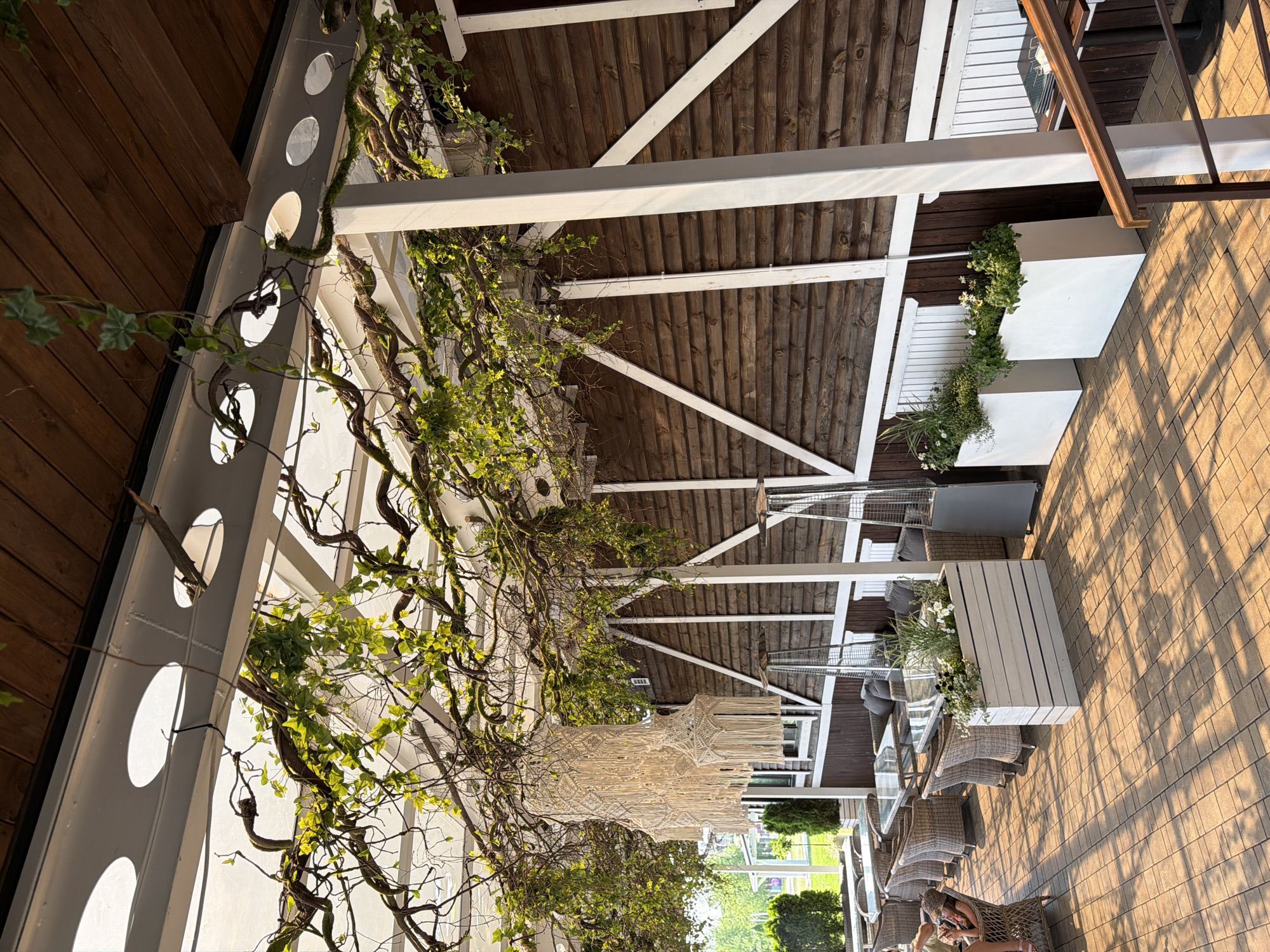 The image depicts an outdoor seating area of a restaurant with a wooden roof structure covered in vines. A macrame chandelier hangs from the ceiling. The seating area features tables and chairs made of wicker, along with several planters containing greenery. Two patio heaters are visible. The floor is paved with interlocking stones. In the background, there are more tables and chairs, and a grassy area with trees. A person wearing a hat is seated in a wicker chair on the left side of the image. The scene is bathed in sunlight, casting shadows across the patio.