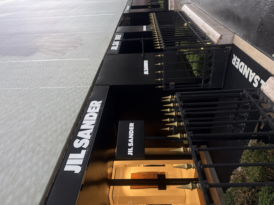 This image depicts the exterior of a Jil Sander boutique in Paris, France, viewed from a slightly elevated perspective on a wet street. The foreground features an ornate black metal fence with numerous gold-colored pointed finials, separating the sidewalk from the store's entrance area. Behind the fence, a dark doormat displaying "JIL SANDER" in white capital letters leads to a glass entrance door. Adjacent to the entrance, a black sign with "JIL SANDER" in white hangs perpendicularly to the building. The store's facade is predominantly black, featuring multiple instances of "JIL SANDER" in raised white capital letters across its length. Through the glass entrance, an illuminated interior is visible, showcasing an abstract, rectangular artwork or display piece with brown and orange tones. Above the storefront, the building is covered with a light-colored, possibly translucent, mesh or scrim material, through which the faint outline of a large human hand is discernible. The street surface is dark and reflective, indicating wet conditions. No individuals are present in the visible scene.