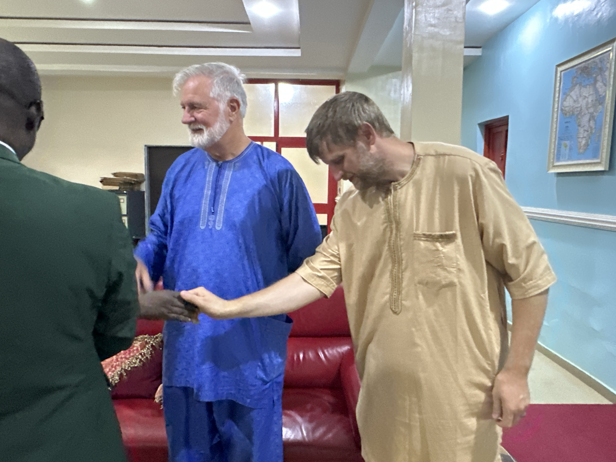 In a brightly lit interior, likely a waiting area or office in Television, Nigeria, three men are engaged in a handshake. On the left, a Black man in a dark green suit is partially visible from the back, his head turned slightly. In the center, an older White man with a gray beard wears a vibrant blue traditional African garment. To his right, another White man, with short brown hair and a beard, is dressed in a tan traditional African outfit. The men are in the process of shaking hands, their faces turned towards each other in a gesture of greeting or farewell. Behind them, a plush red leather sofa is visible, suggesting a comfortable seating area. The room's walls are painted in light cream and a striking sky blue, with a framed map of Africa hanging on the blue wall. The ceiling features modern recessed lighting. The overall impression is one of a formal yet welcoming environment, possibly a governmental or diplomatic setting.